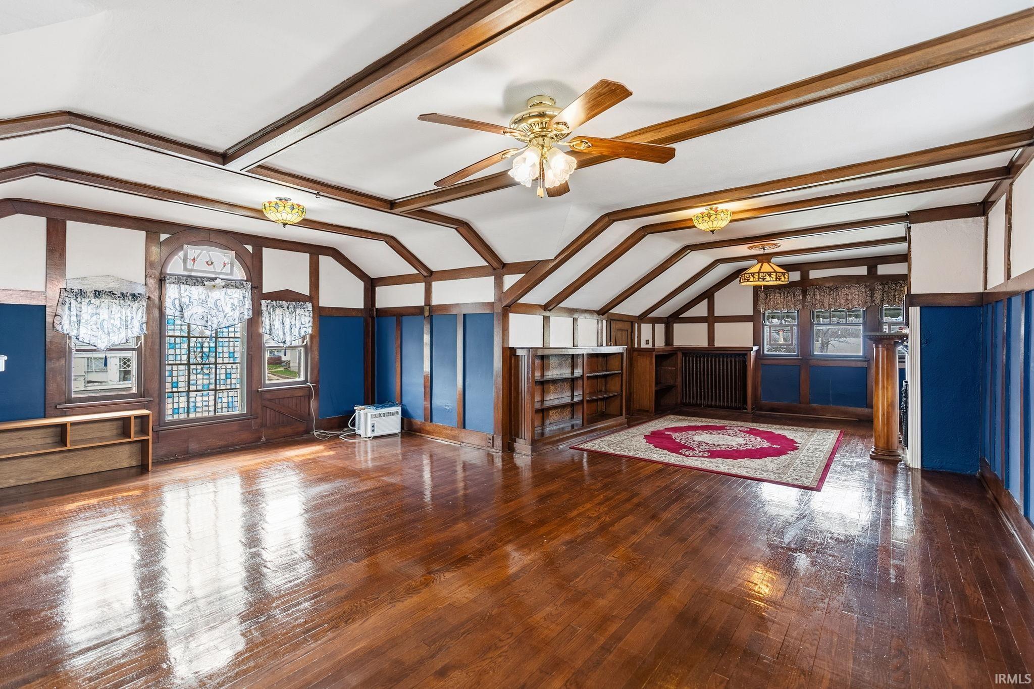 Unfurnished living room featuring dark wood-style flooring, lofted ceiling with beams, a ceiling fan, and radiator