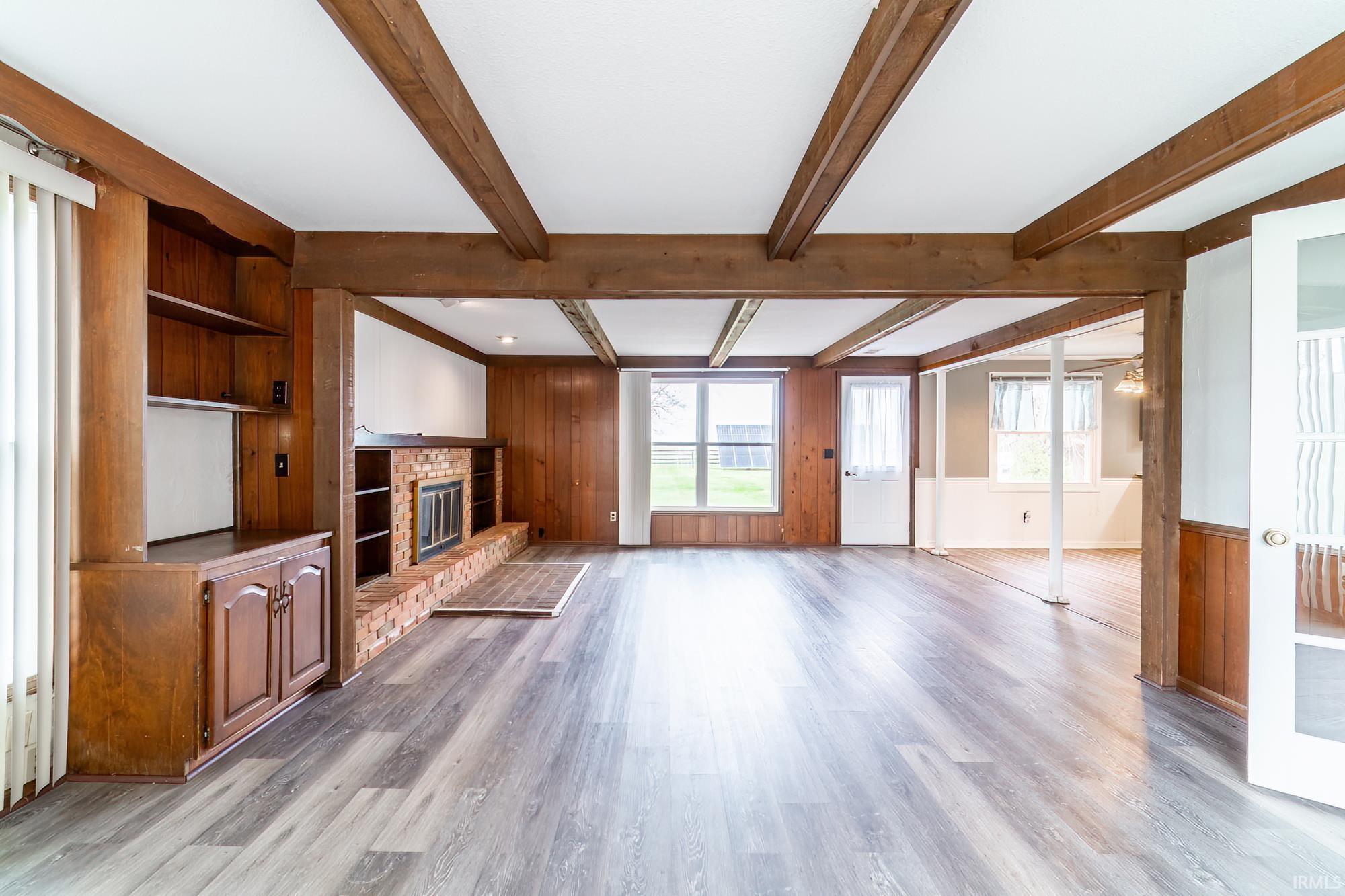 Unfurnished living room featuring wooden walls, a brick fireplace, beamed ceiling, light wood-style flooring, and wainscoting