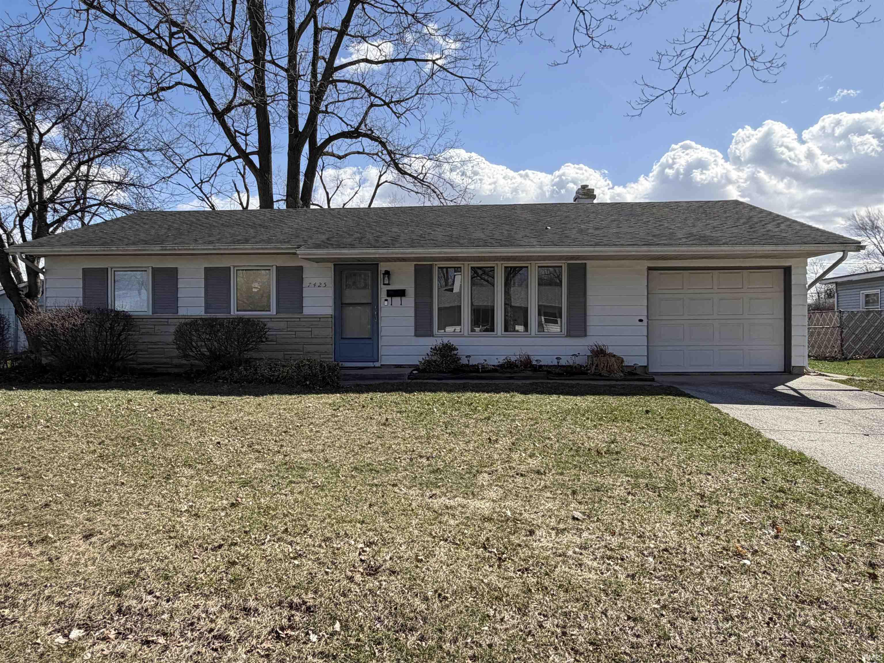 Ranch-style home featuring an attached garage, driveway, a front lawn, a chimney, and roof with shingles