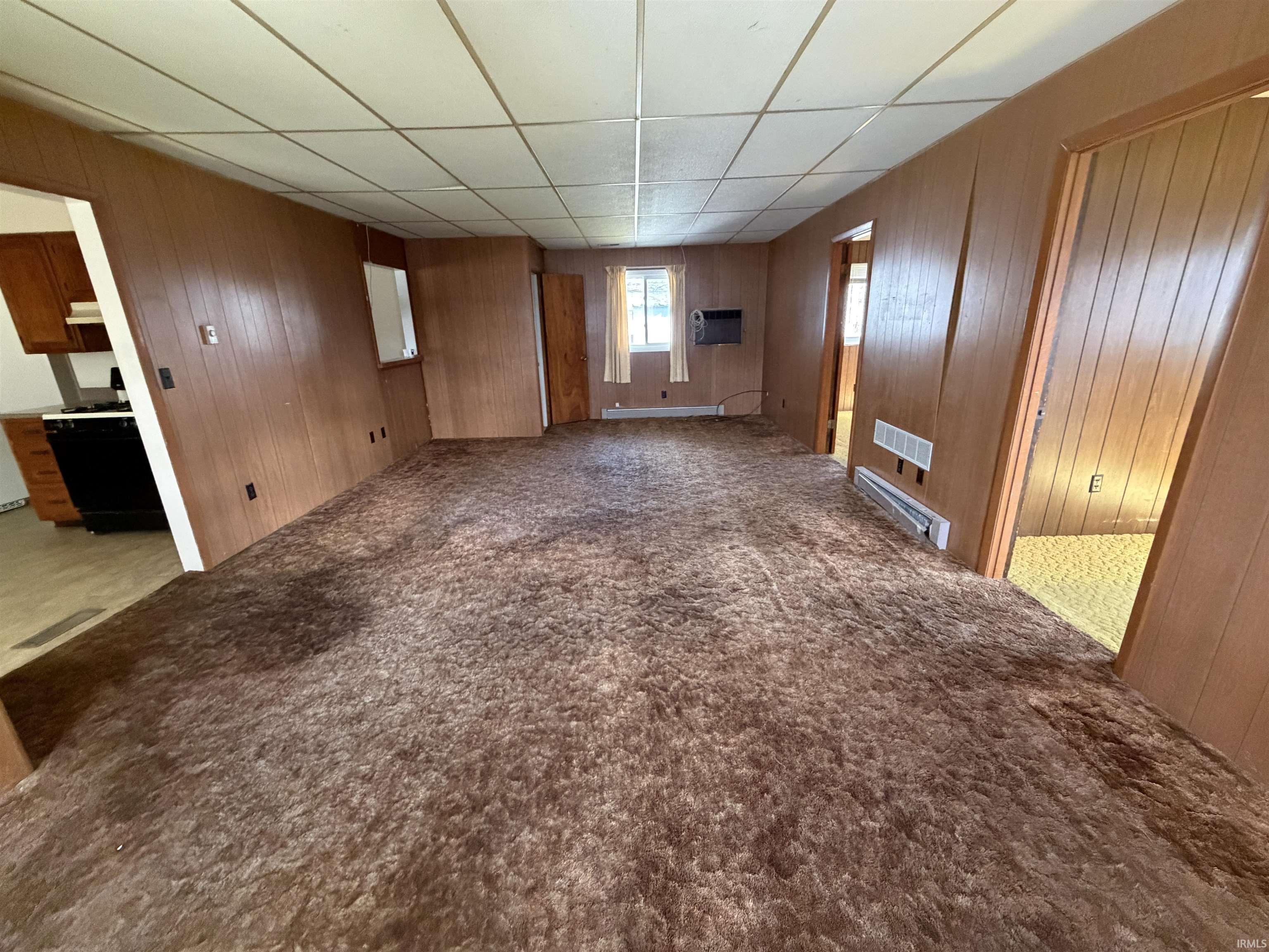 Empty room with carpet flooring, baseboard heating, wood walls, and a drop ceiling