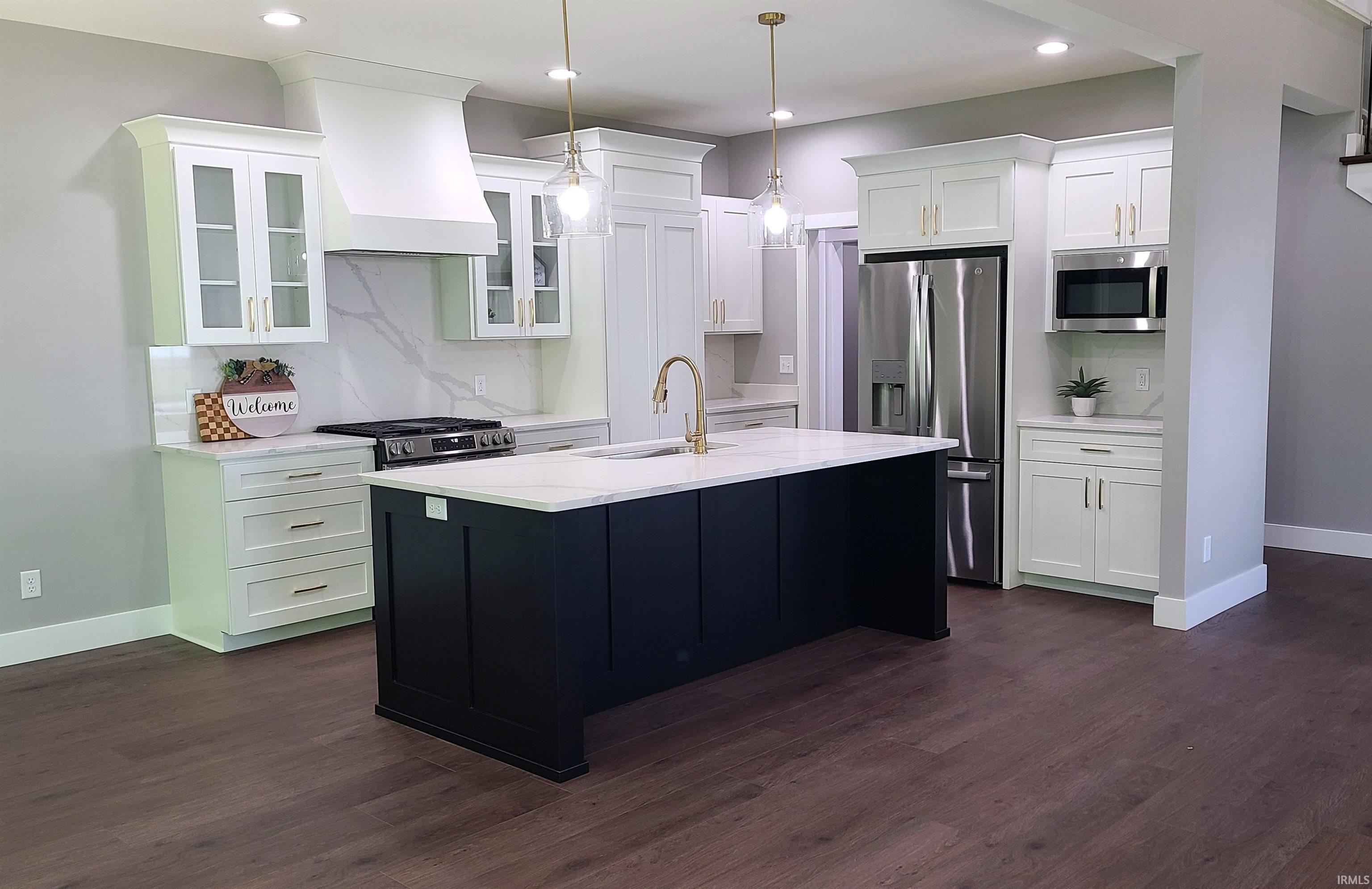 Two tone kitchen with stainless steel appliances, an island with sink, two tone cabinetry, light stone counters, and dark wood-style flooring