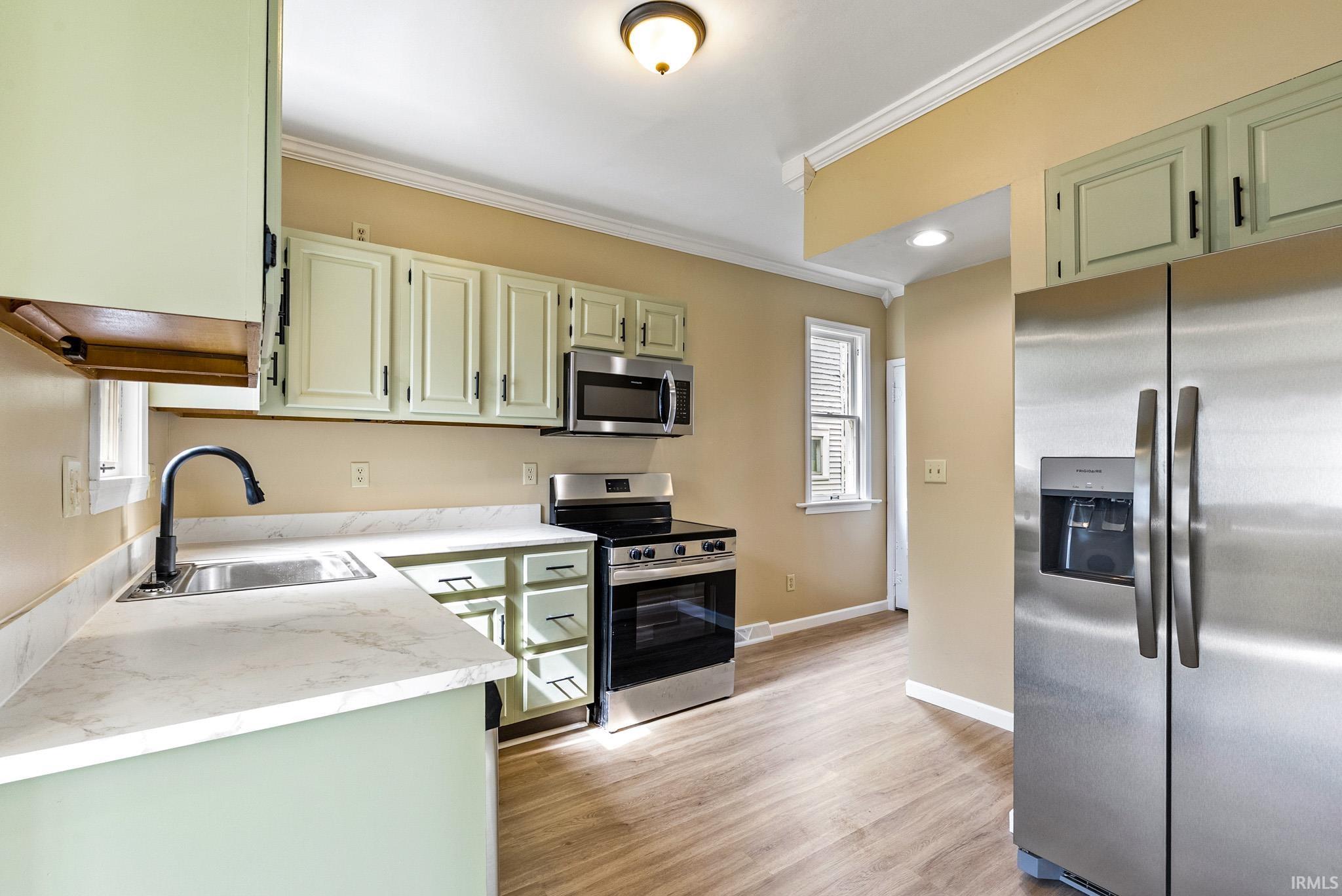 Kitchen with green cabinetry, stainless steel appliances, ornamental molding, light countertops, and light wood-style flooring