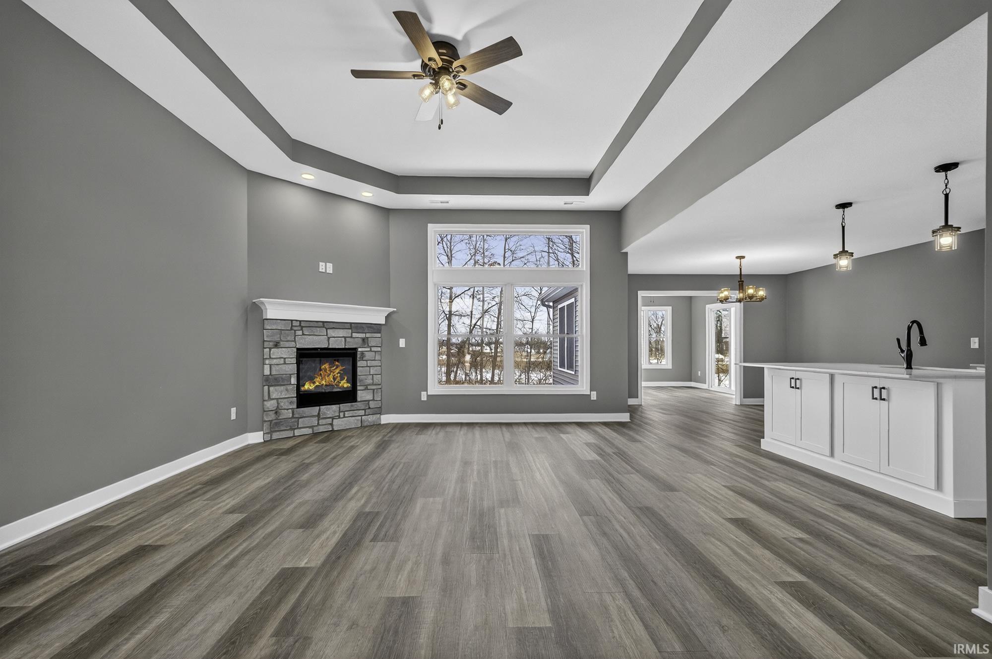 Unfurnished living room with a stone fireplace, a raised ceiling, ceiling fan, dark wood-style flooring, and suspended lighting