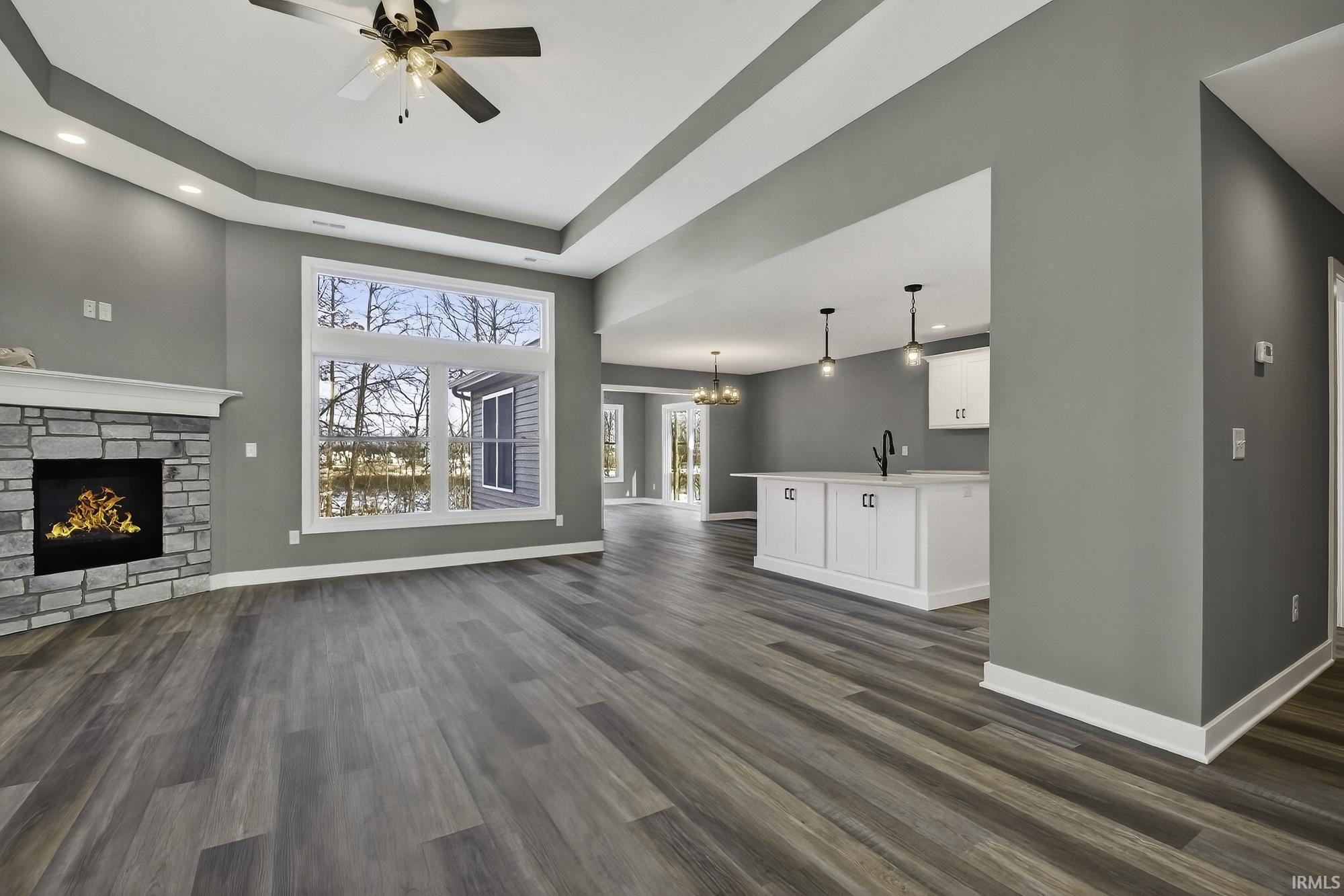 Unfurnished living room featuring a stone fireplace, ceiling fan, a tray ceiling, dark wood-style flooring, and a chandelier