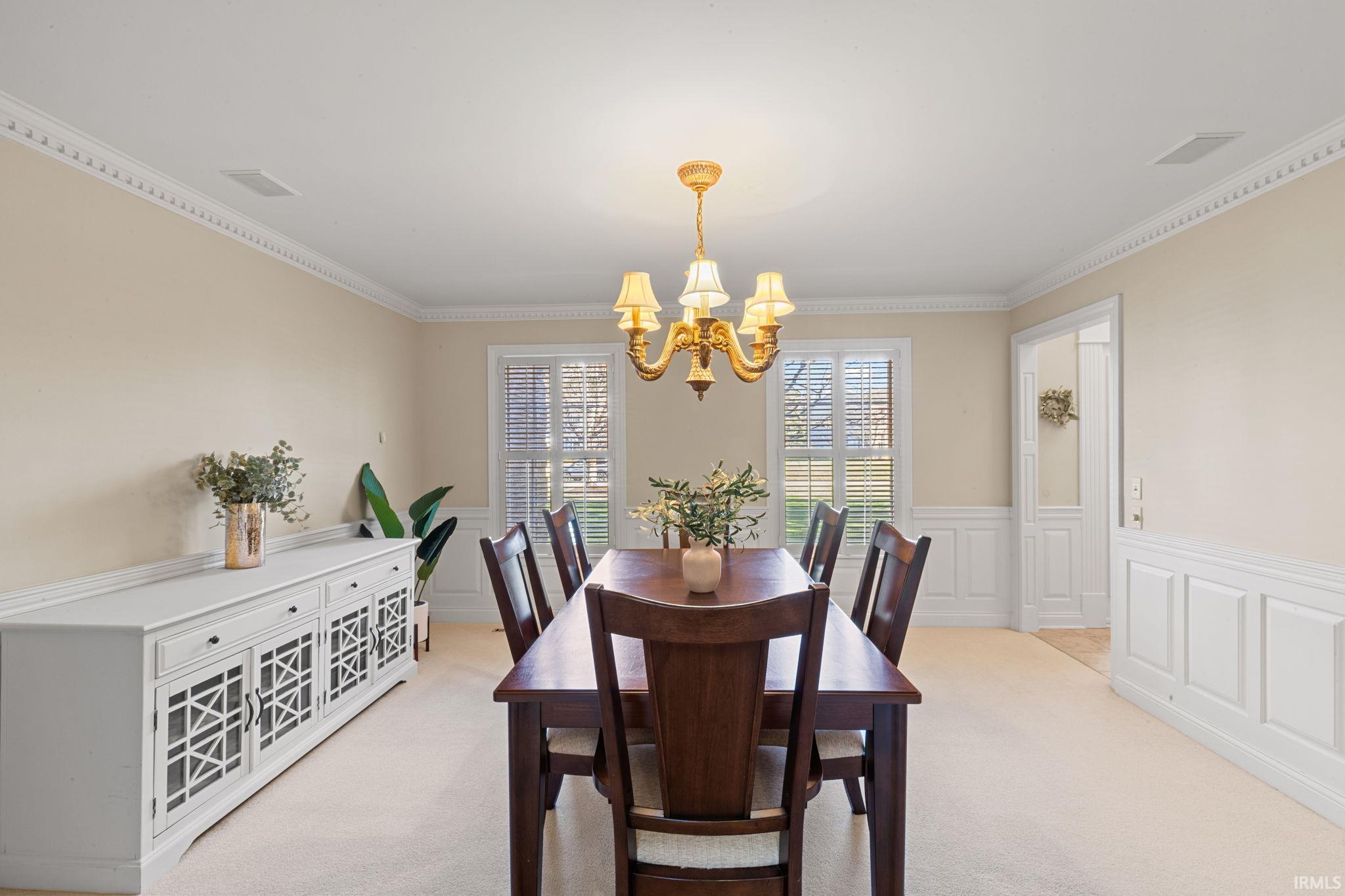 Dining space featuring hanging lights, a decorative wall, wainscoting, light colored carpet, and crown molding