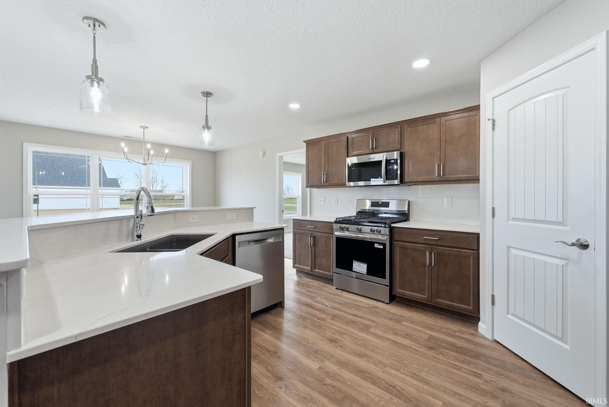 Kitchen with stainless steel appliances, dark wood finish cabinetry, light wood finished floors, hanging lights, and light stone counters