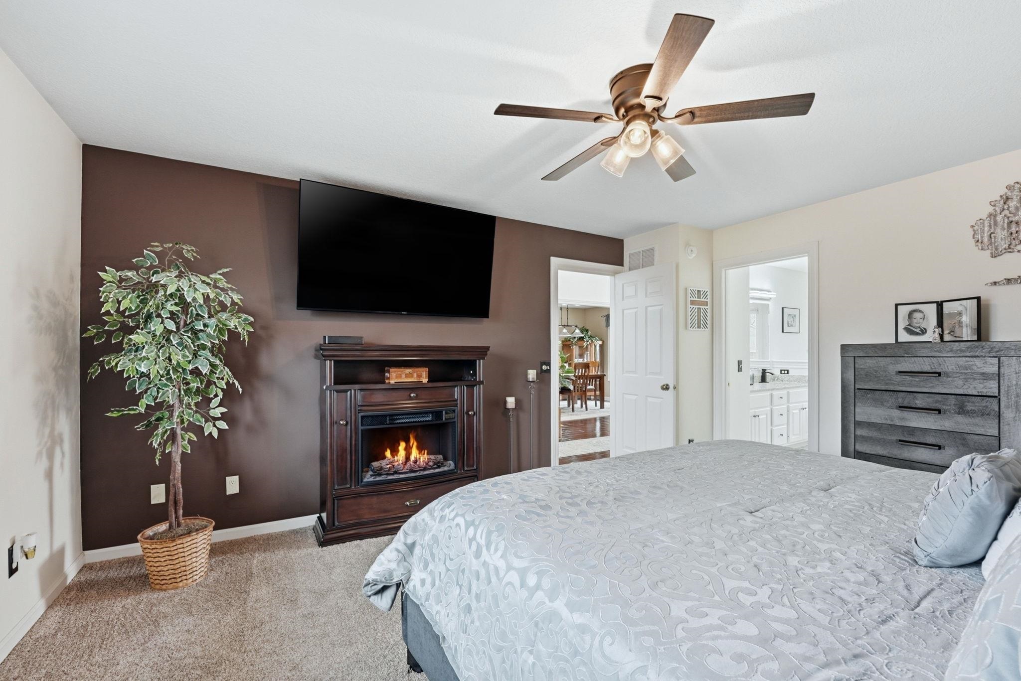 Bedroom with light carpet, a ceiling fan, a lit fireplace, and connected bathroom