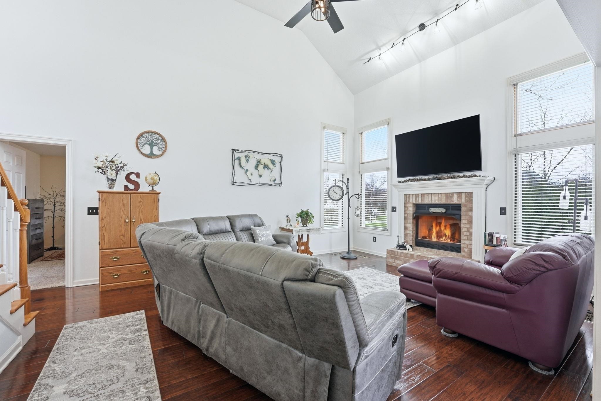 Living room with rail lighting, a ceiling fan, dark wood-style floors, vaulted ceiling, and a brick fireplace