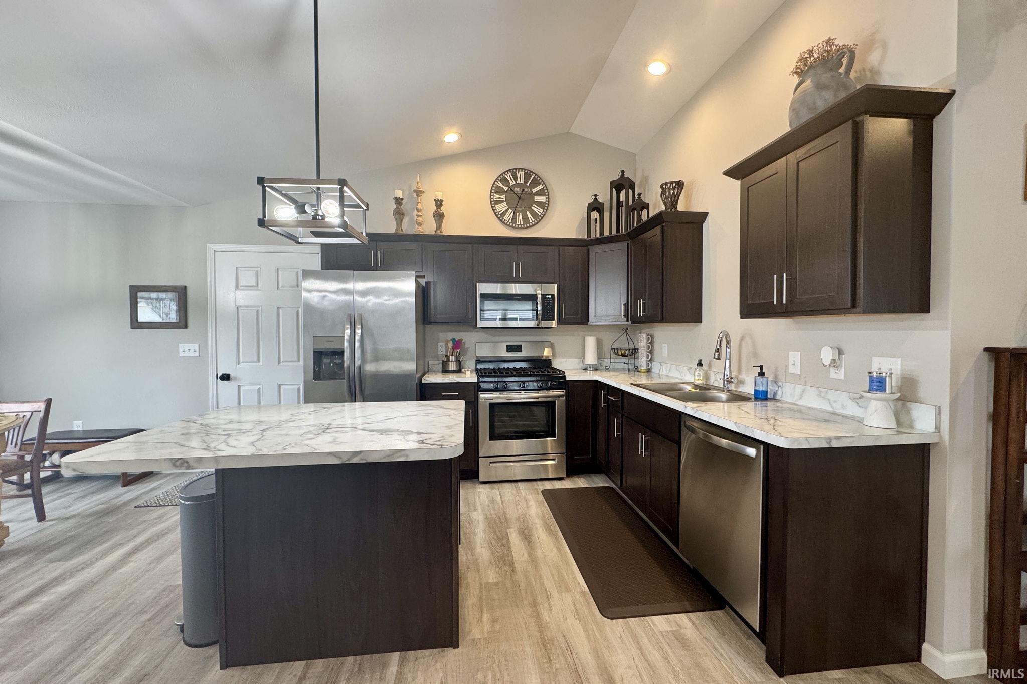 Kitchen with dark wood finish cabinetry, stainless steel appliances, light countertops, vaulted ceiling, and light wood-style floors