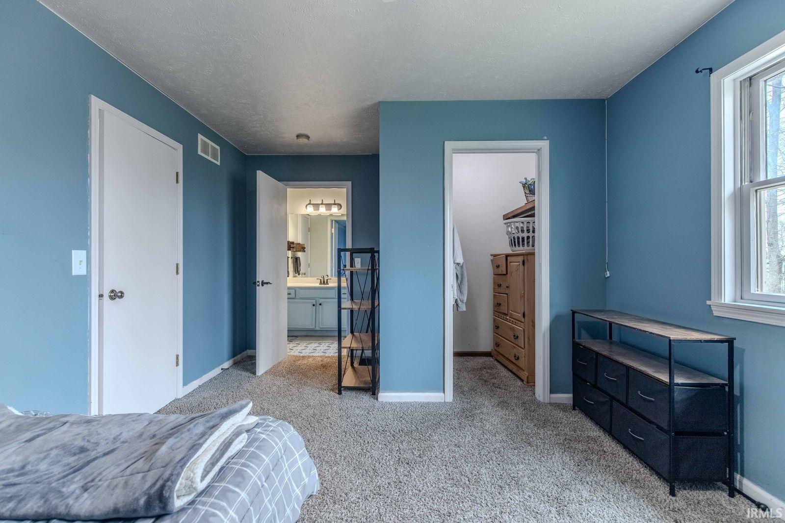 Bedroom with a walk in closet, light colored carpet, multiple windows, ensuite bathroom, and a textured ceiling