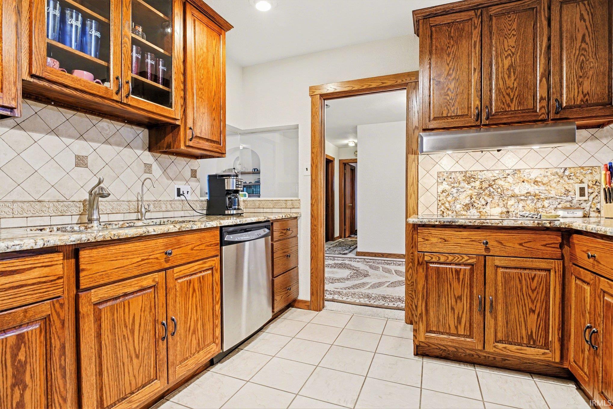 Kitchen with glass insert cabinets, light stone counters, wood finish cabinets, stainless steel dishwasher, and exhaust hood