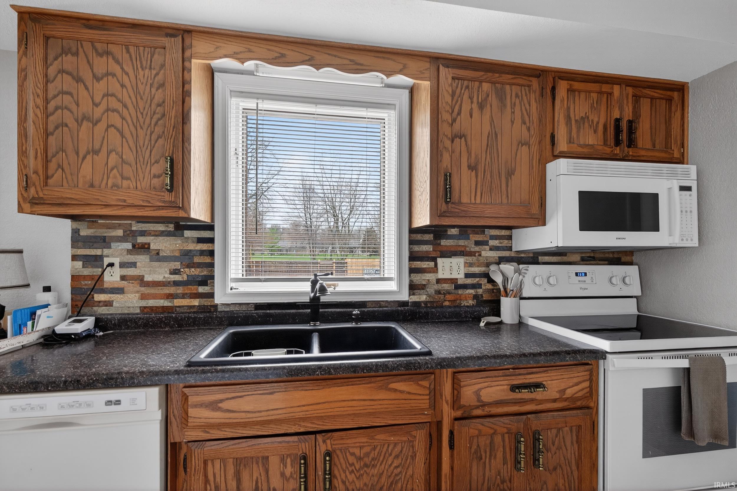 Kitchen with white appliances, dark countertops, wood finish cabinets, and backsplash