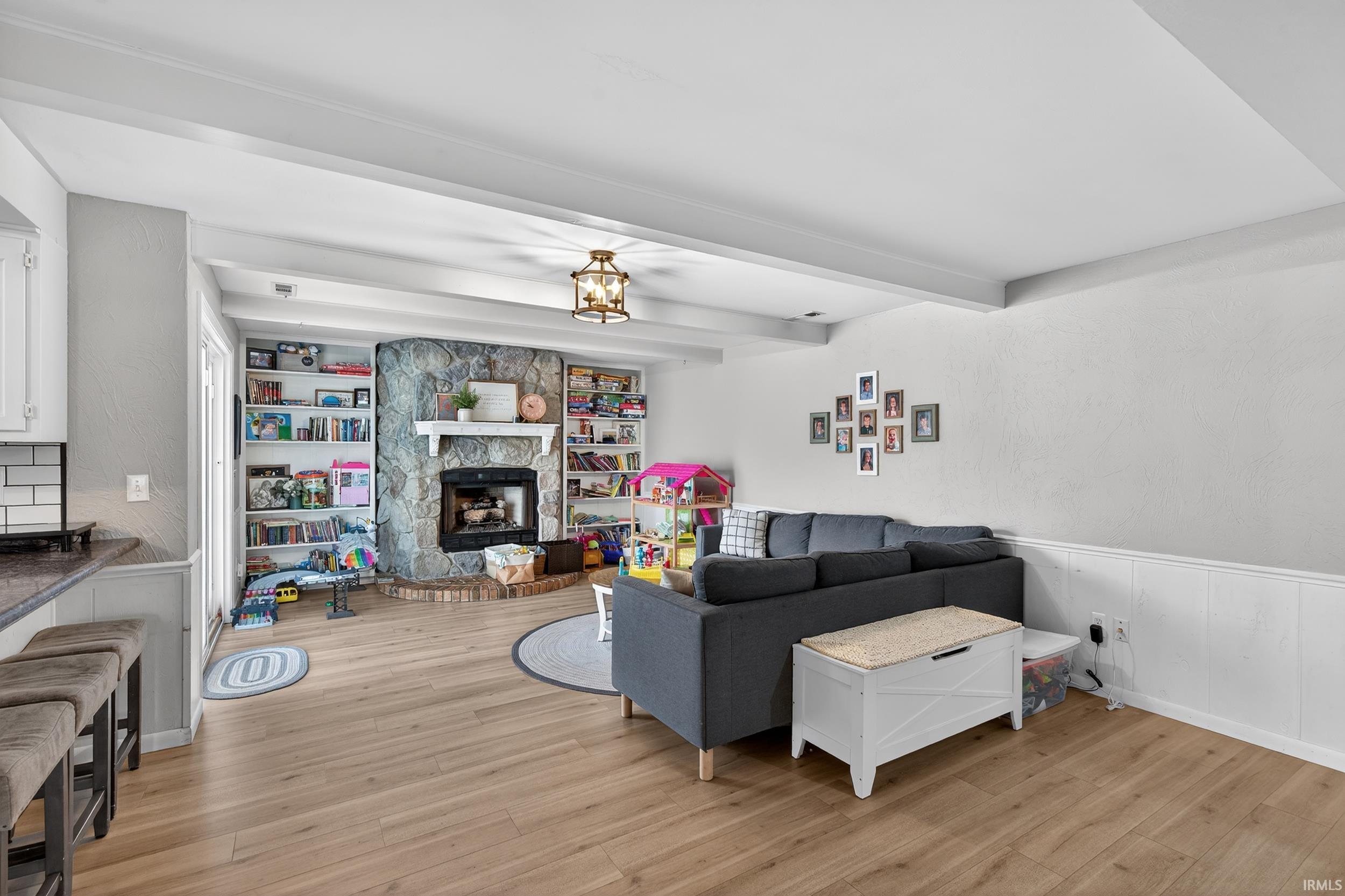 Living room featuring light wood-style floors, wainscoting, a stone fireplace, and beamed ceiling