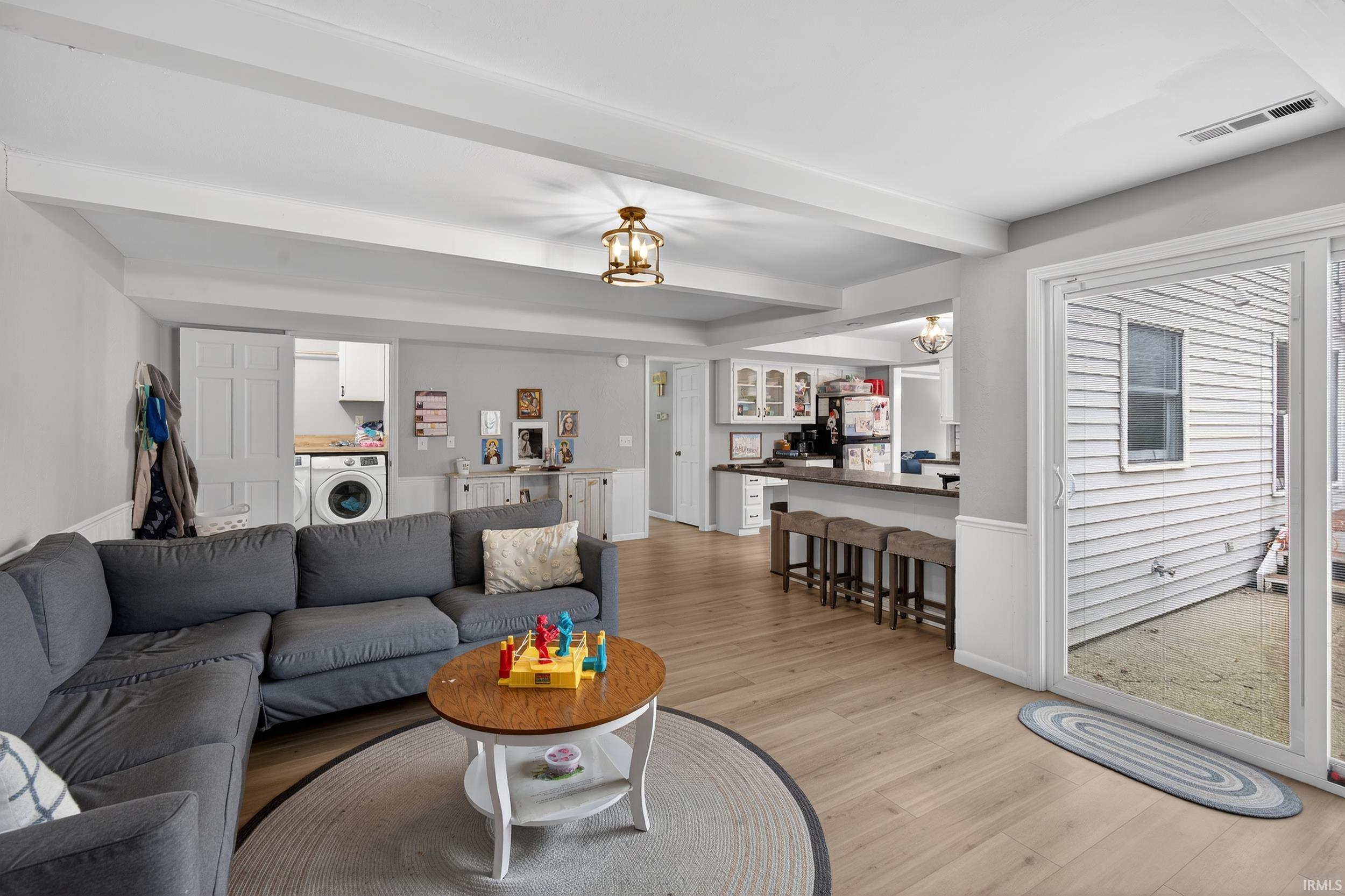 Living room with light wood finished floors, washer / dryer, wainscoting, a chandelier, and beamed ceiling