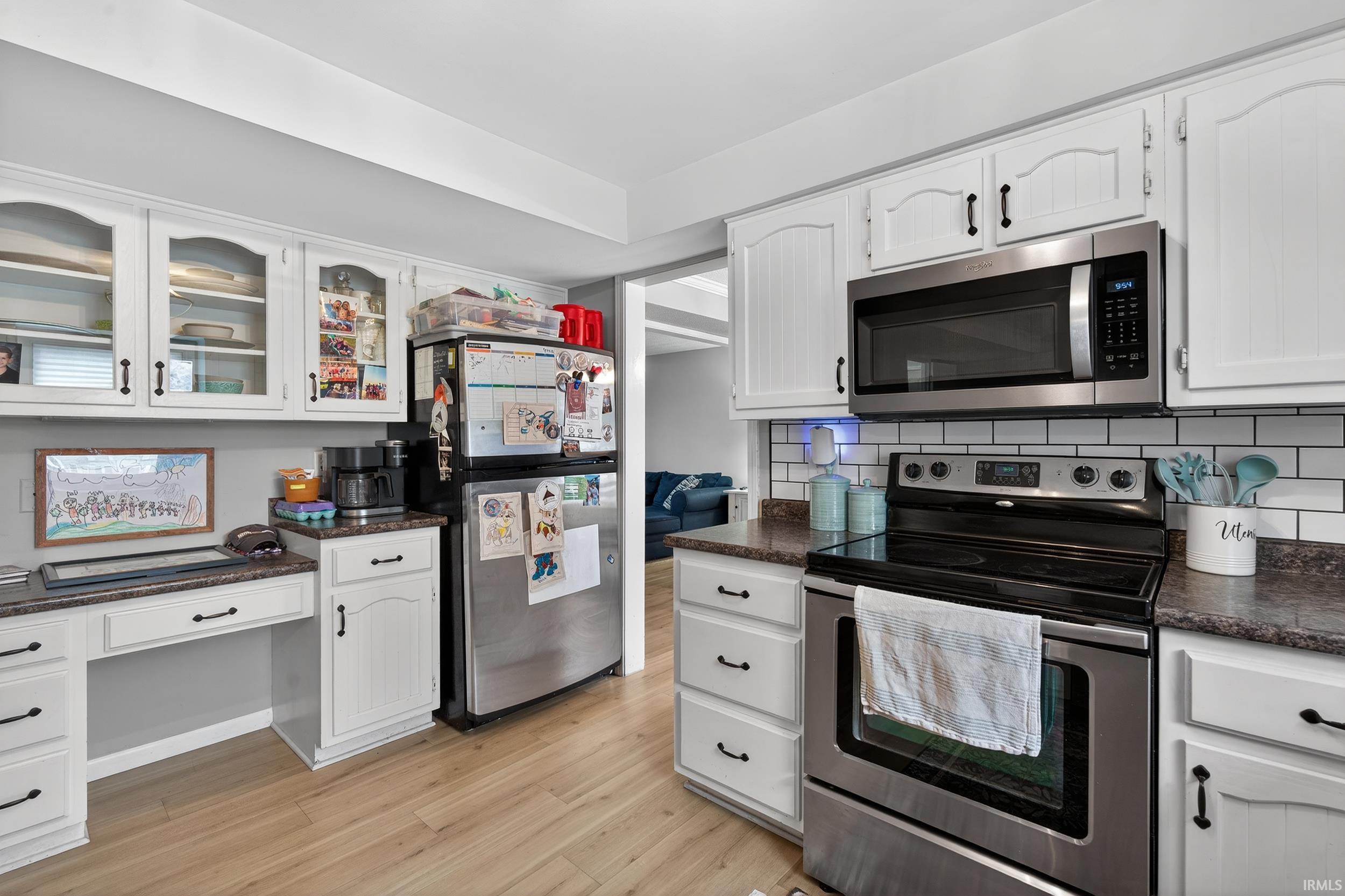 Kitchen featuring stainless steel appliances, white cabinets, dark countertops, light wood-style floors, and glass fronted cabinets