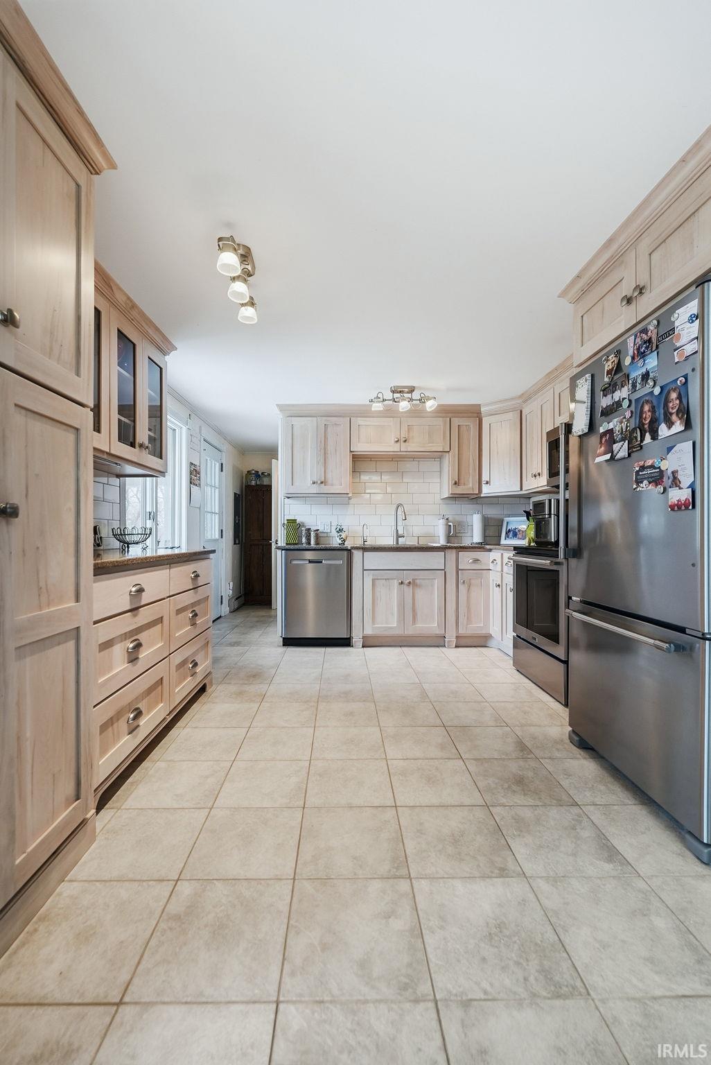 Kitchen featuring light wood finish cabinetry, stainless steel appliances, tasteful backsplash, light tile patterned flooring, and glass insert cabinets