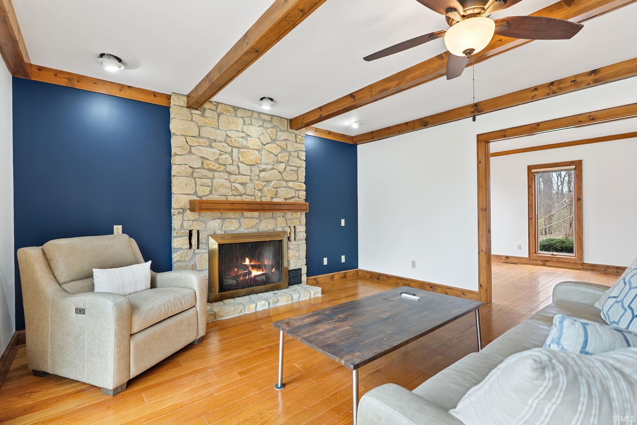 Living area with a stone fireplace, light wood-style flooring, a ceiling fan, and beam ceiling