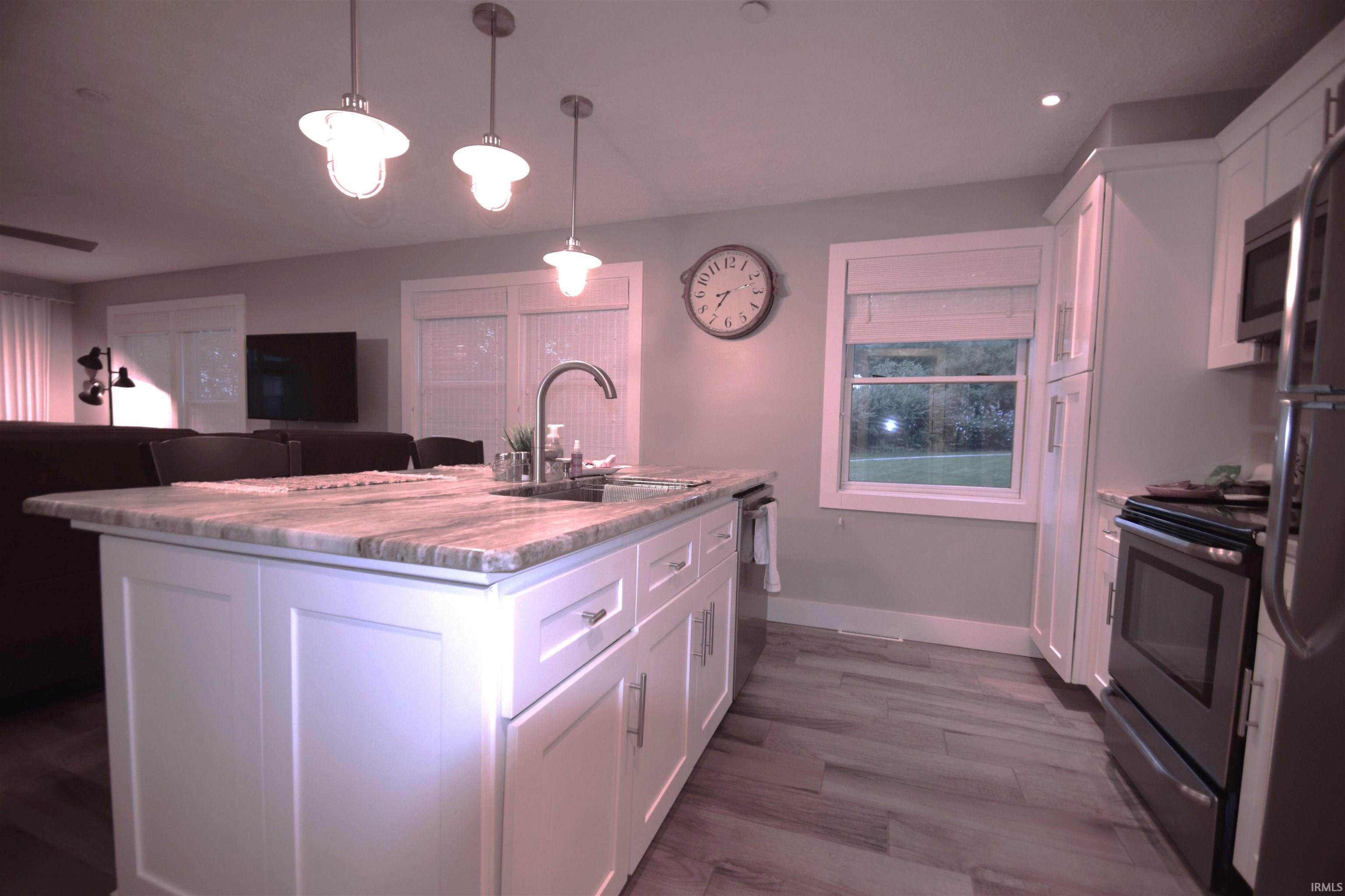 Kitchen featuring white cabinets, stainless steel appliances, a center island with sink, decorative light fixtures, and light stone counters
