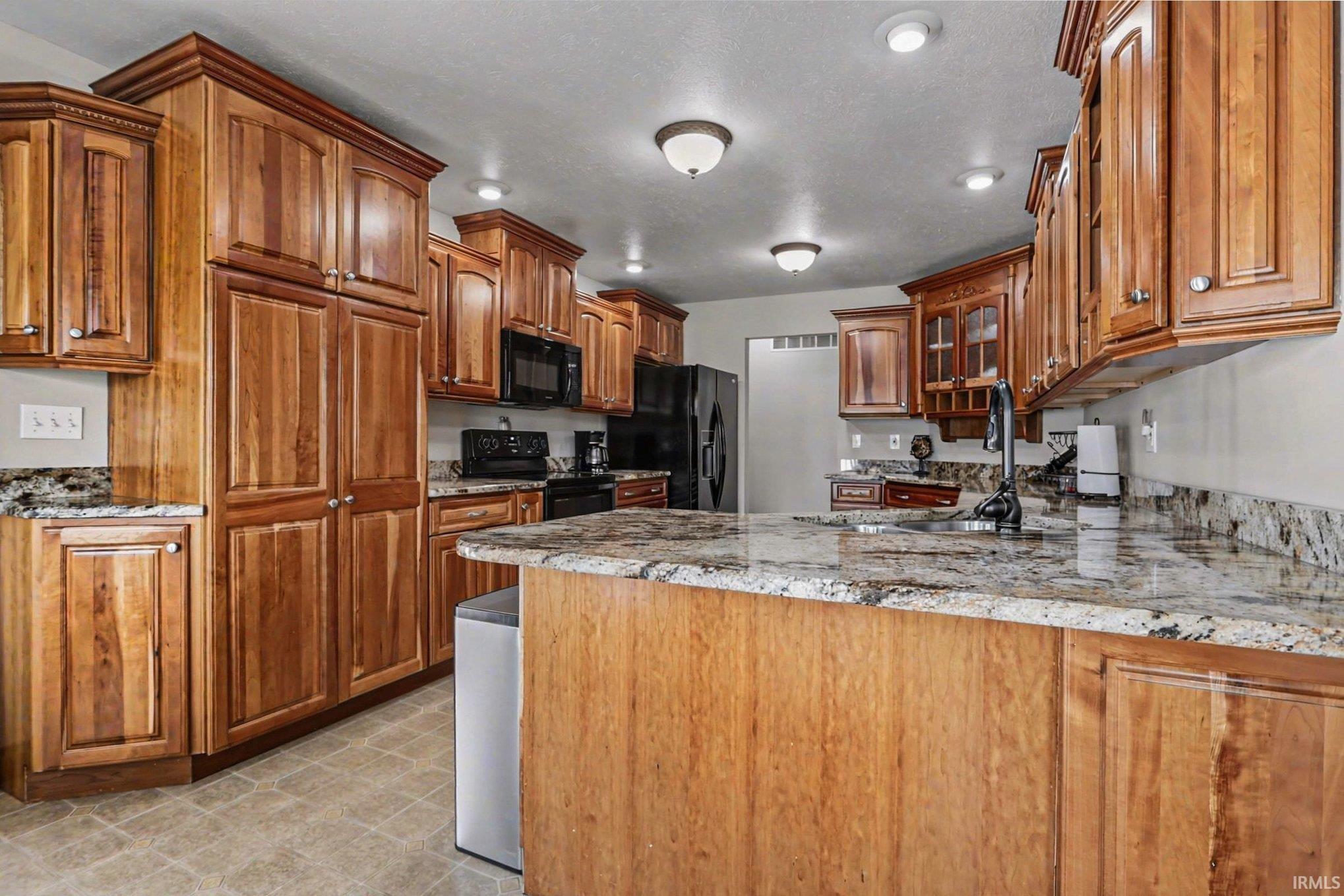 Kitchen with wood finish cabinetry, black appliances, a peninsula, light floors, and light stone counters