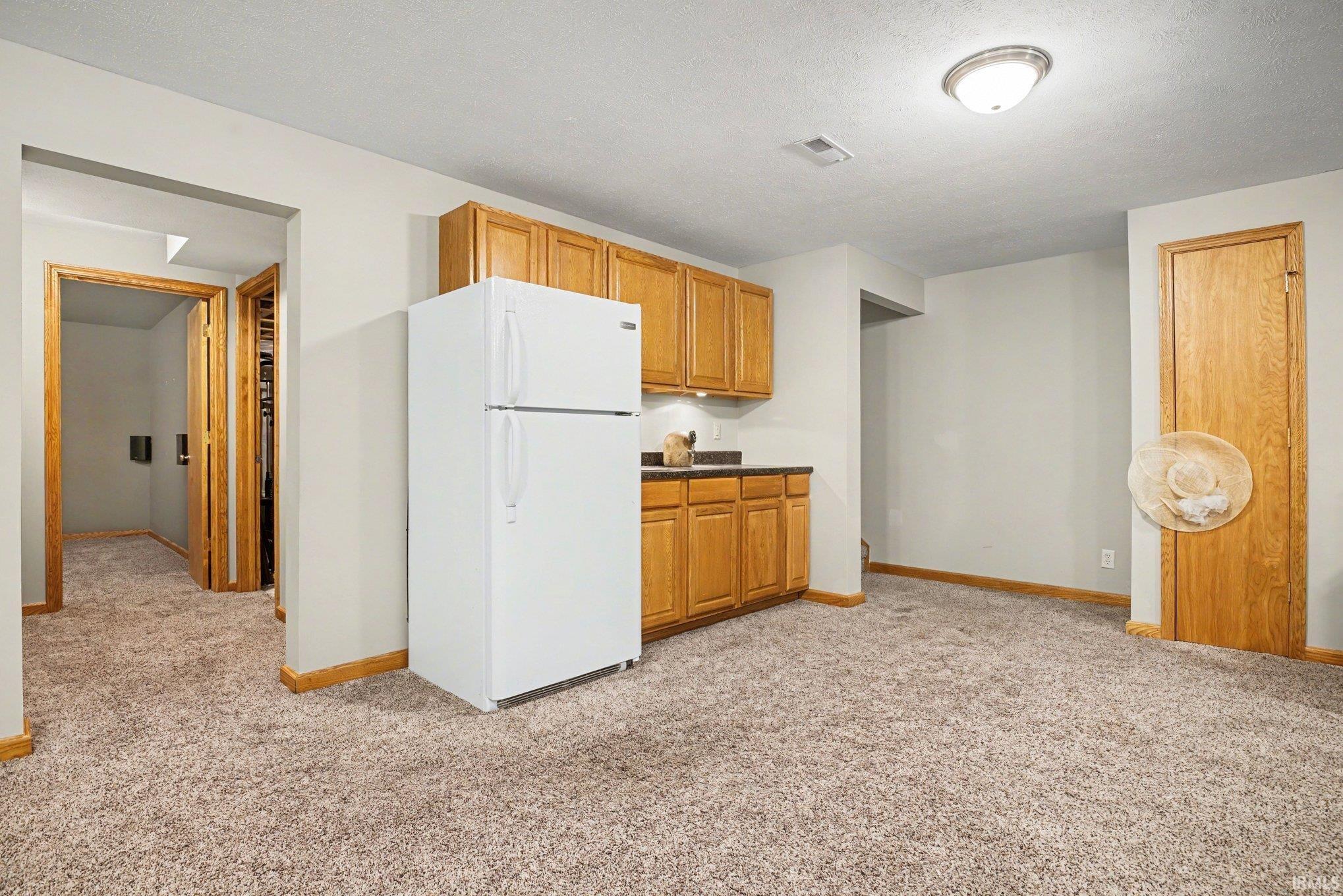Kitchen with freestanding refrigerator, light carpet, a textured ceiling, wood finish cabinetry, and dark countertops