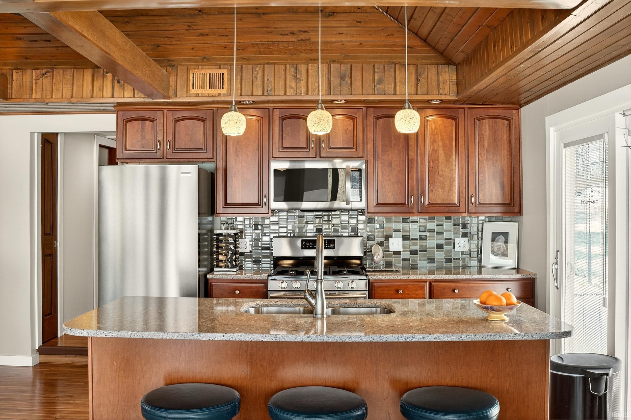 Kitchen featuring light stone counters, a vaulted wooden ceiling, a kitchen island with sink, stainless steel appliances, and pendant lighting
