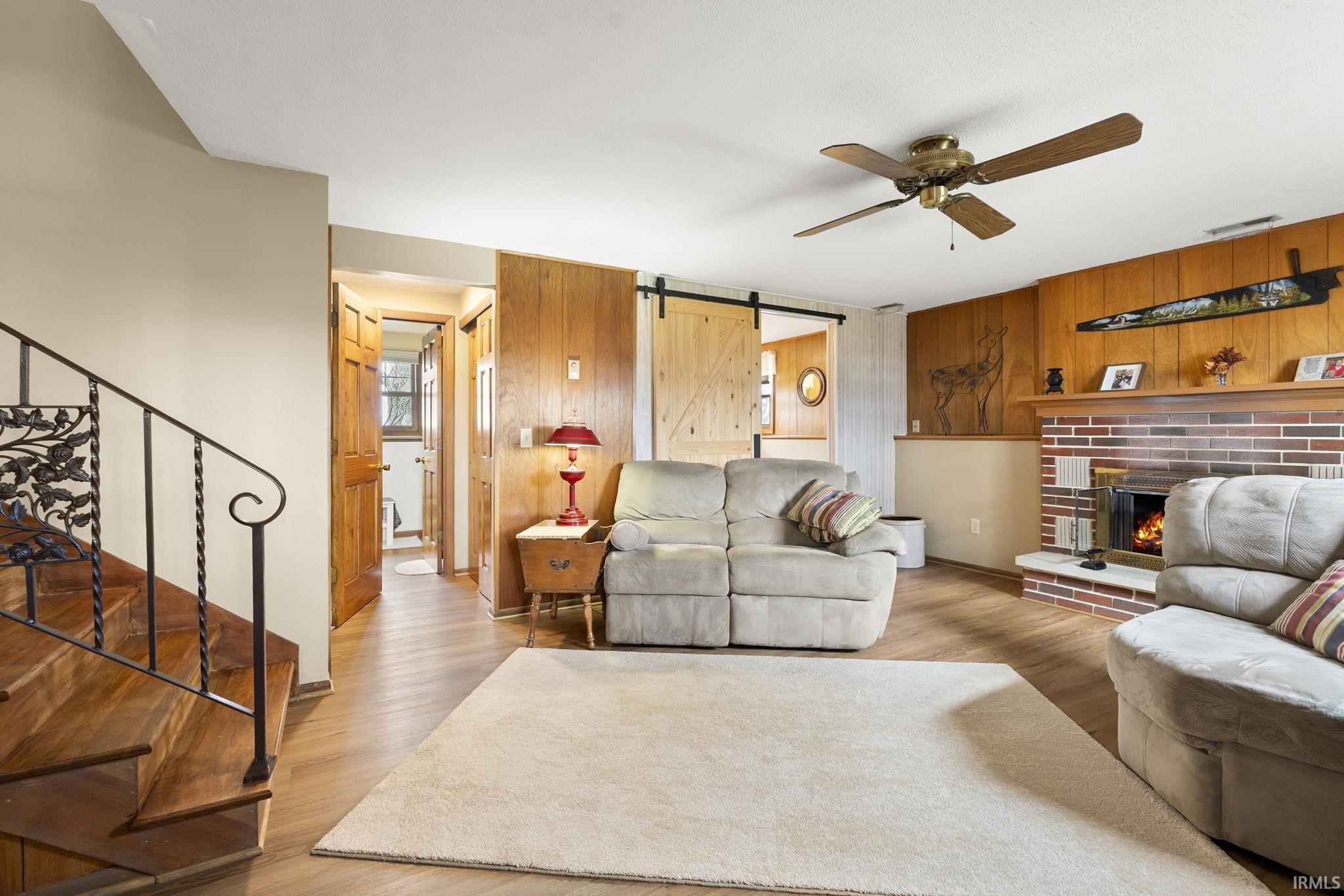 Living room with a barn door, a ceiling fan, wood finished floors, a fireplace, and wooden walls