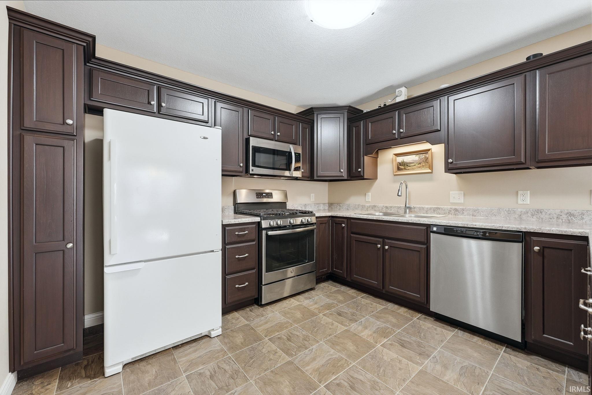 Kitchen featuring stainless steel appliances, dark wood finish cabinets, stone finish flooring, and light stone countertops