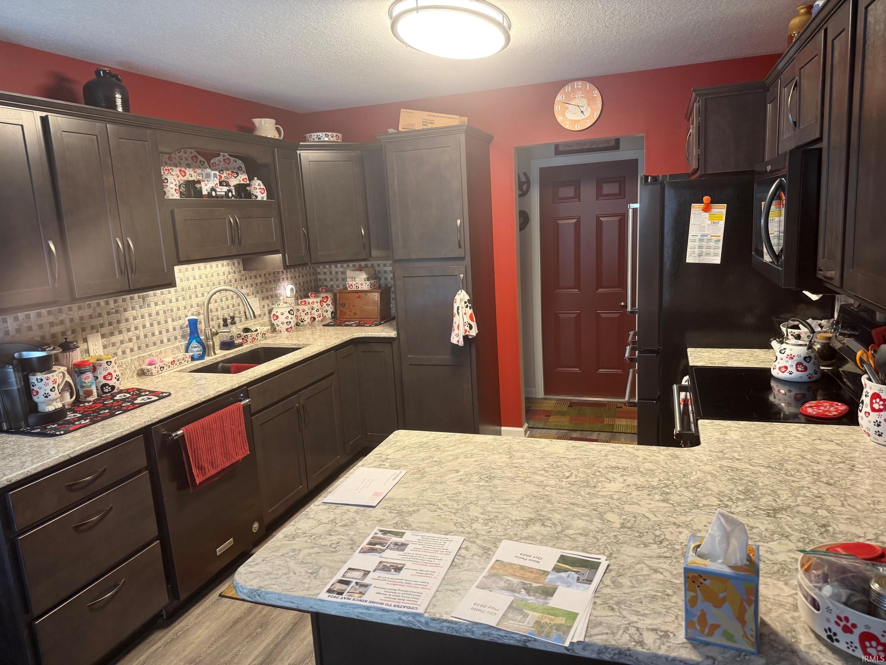 Kitchen with dark wood finish cabinets, black appliances, a textured ceiling, backsplash, and light stone counters