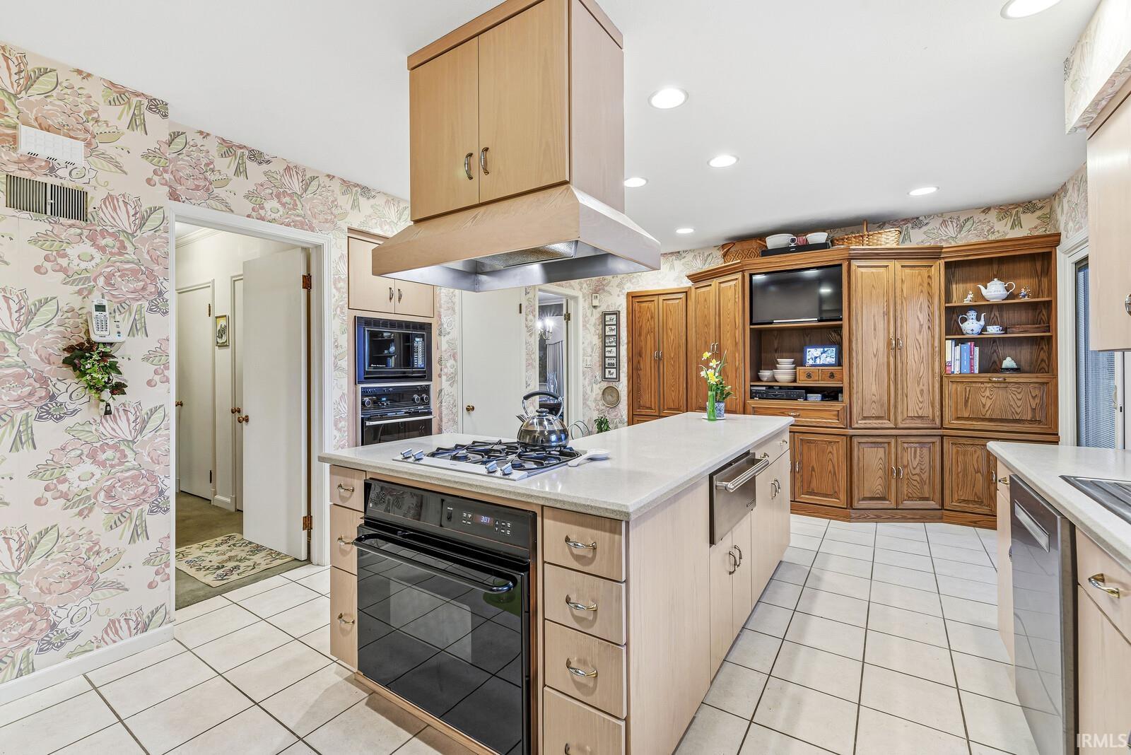 Dual tone kitchen featuring wallpapered walls, black appliances, two tone cabinets, light tile patterned floors, and a center island