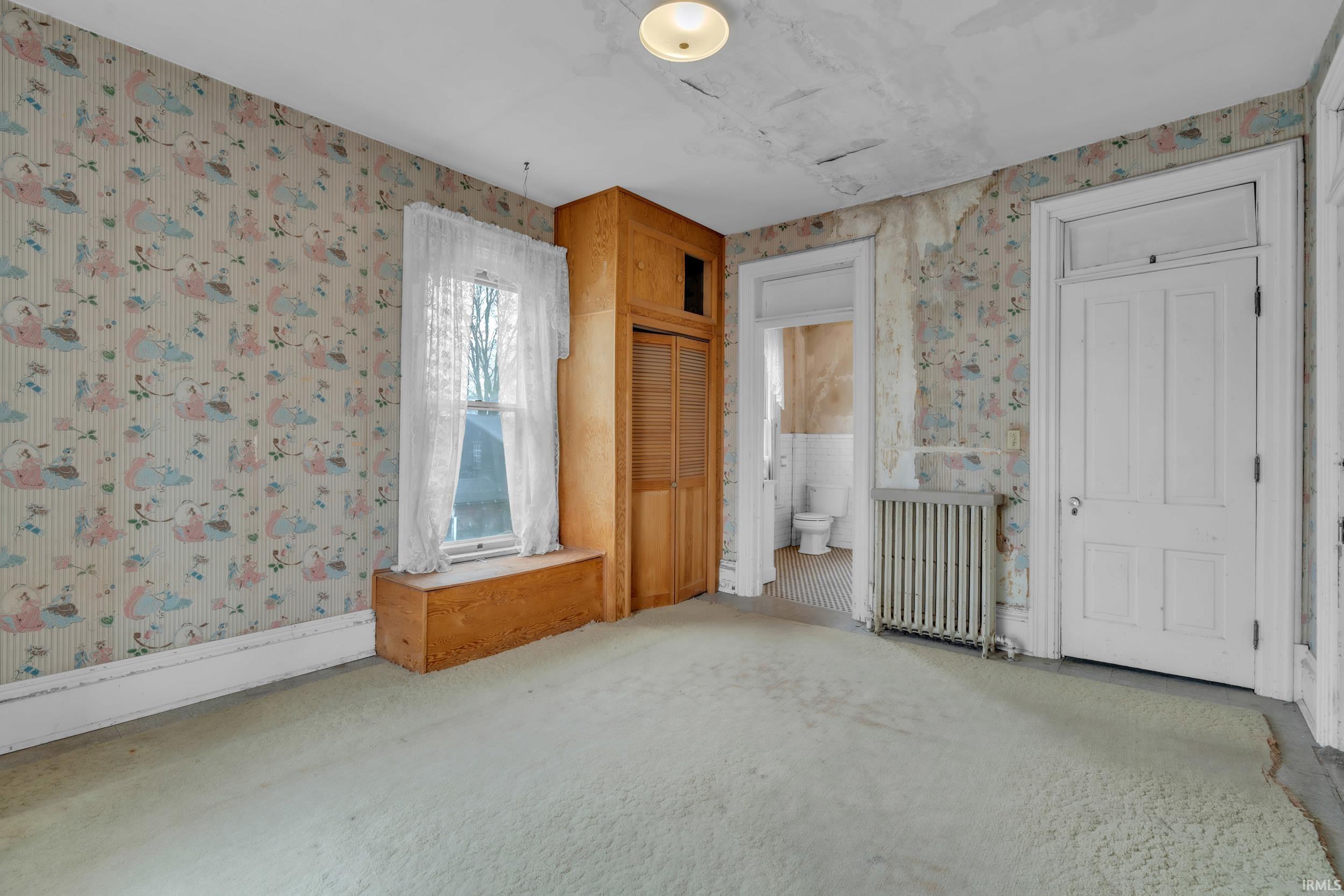 Unfurnished bedroom with wallpapered walls, radiator, carpet flooring, ensuite bathroom, and baseboard heating