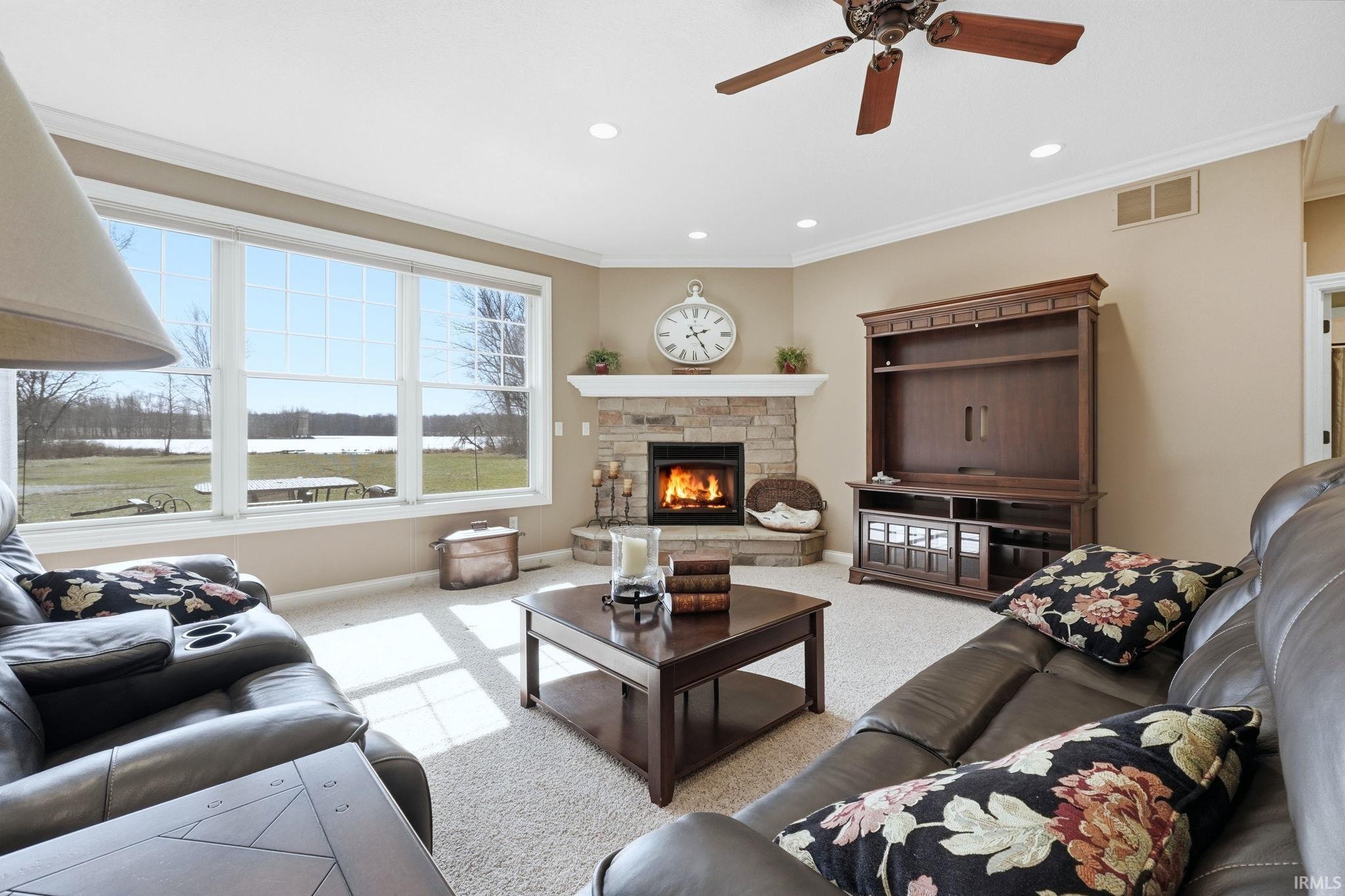 Living room featuring a stone fireplace, light colored carpet, a ceiling fan, ornamental molding, and recessed lighting