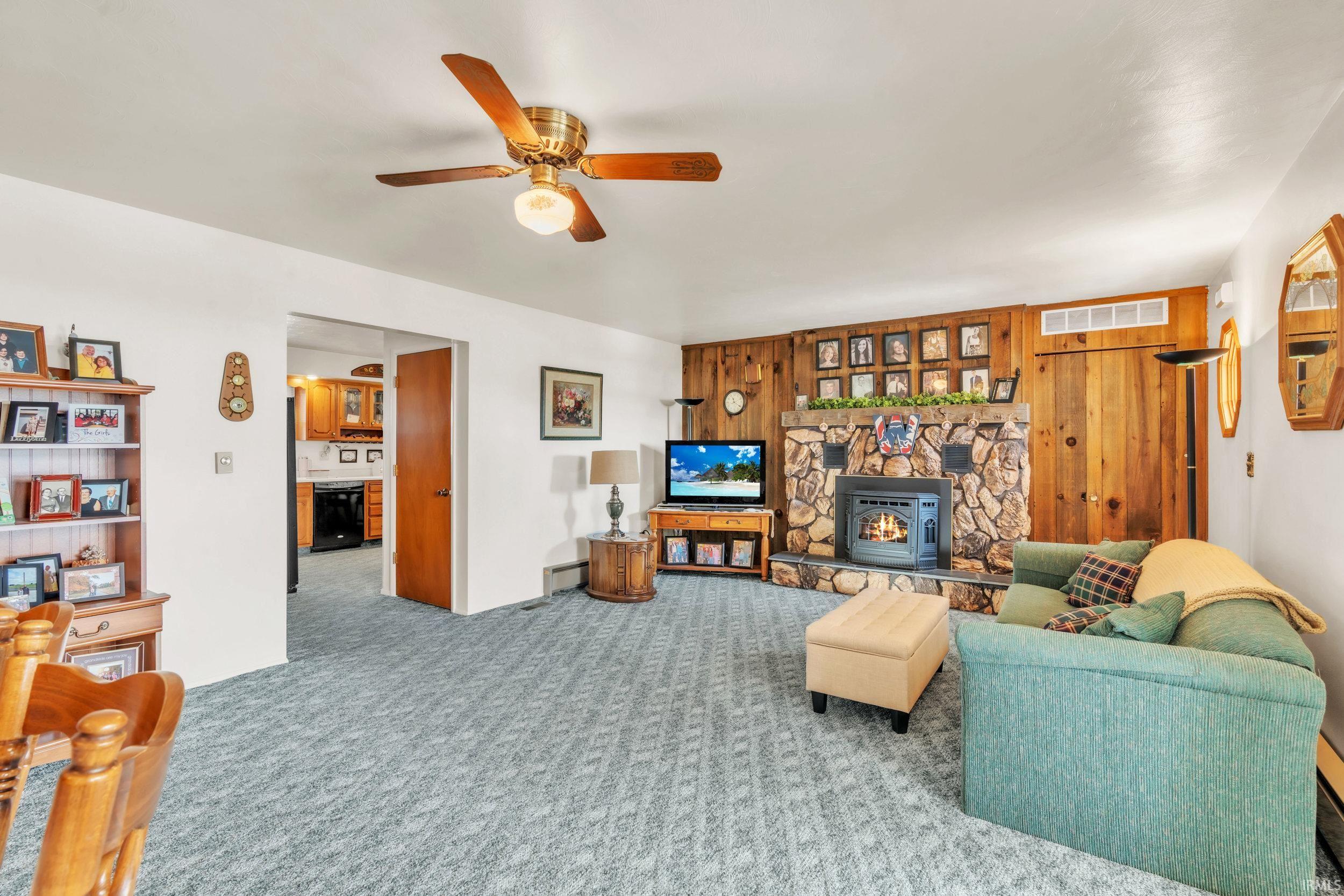 Living room featuring a ceiling fan, carpet floors, a wood stove, wooden walls, and a baseboard radiator