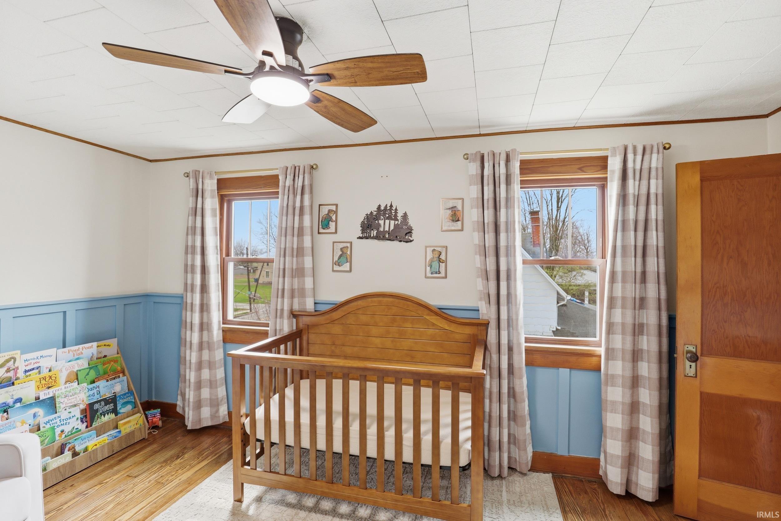 Bedroom featuring light wood-type flooring, crown molding, a nursery area, ceiling fan, and a wainscoted wall