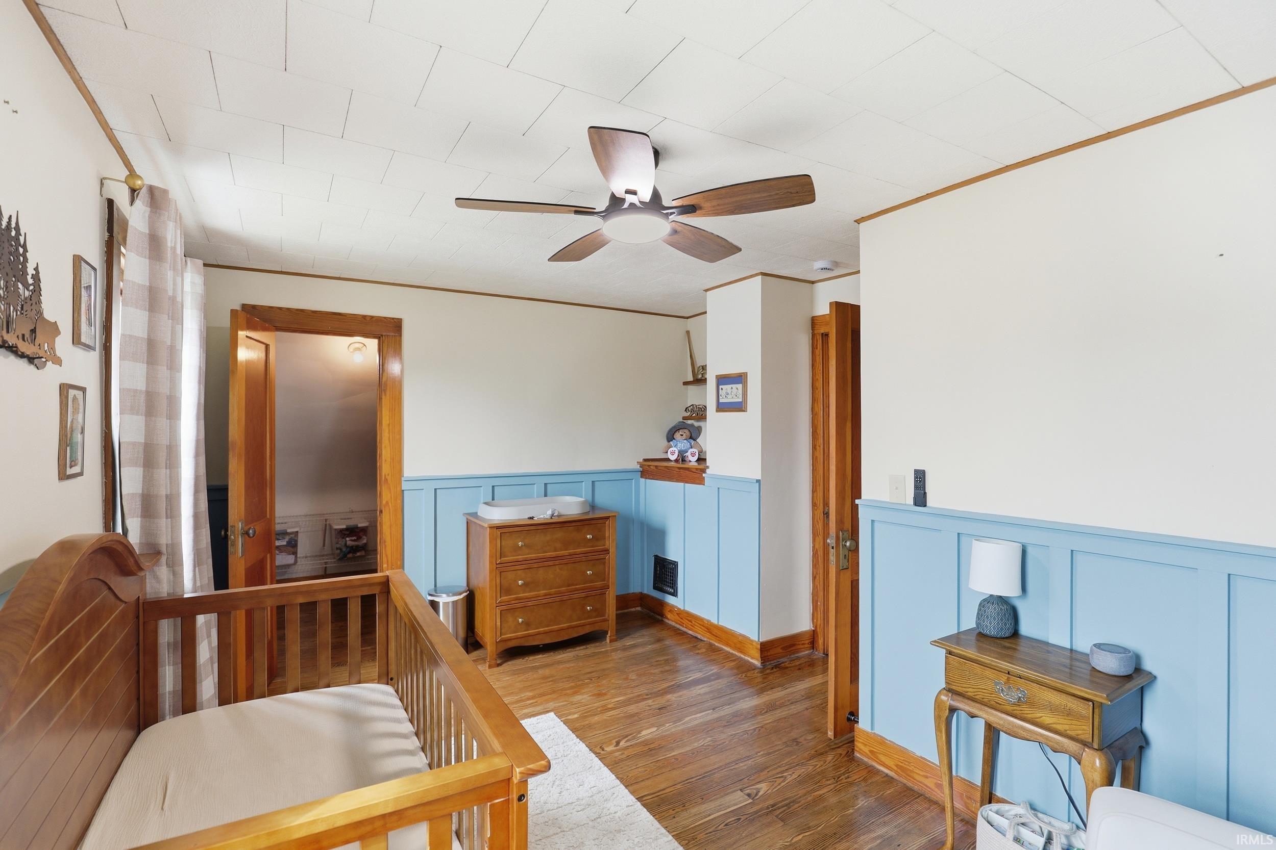 Bedroom featuring wood-type flooring, ornamental molding, a wainscoted wall, ceiling fan, and a decorative wall