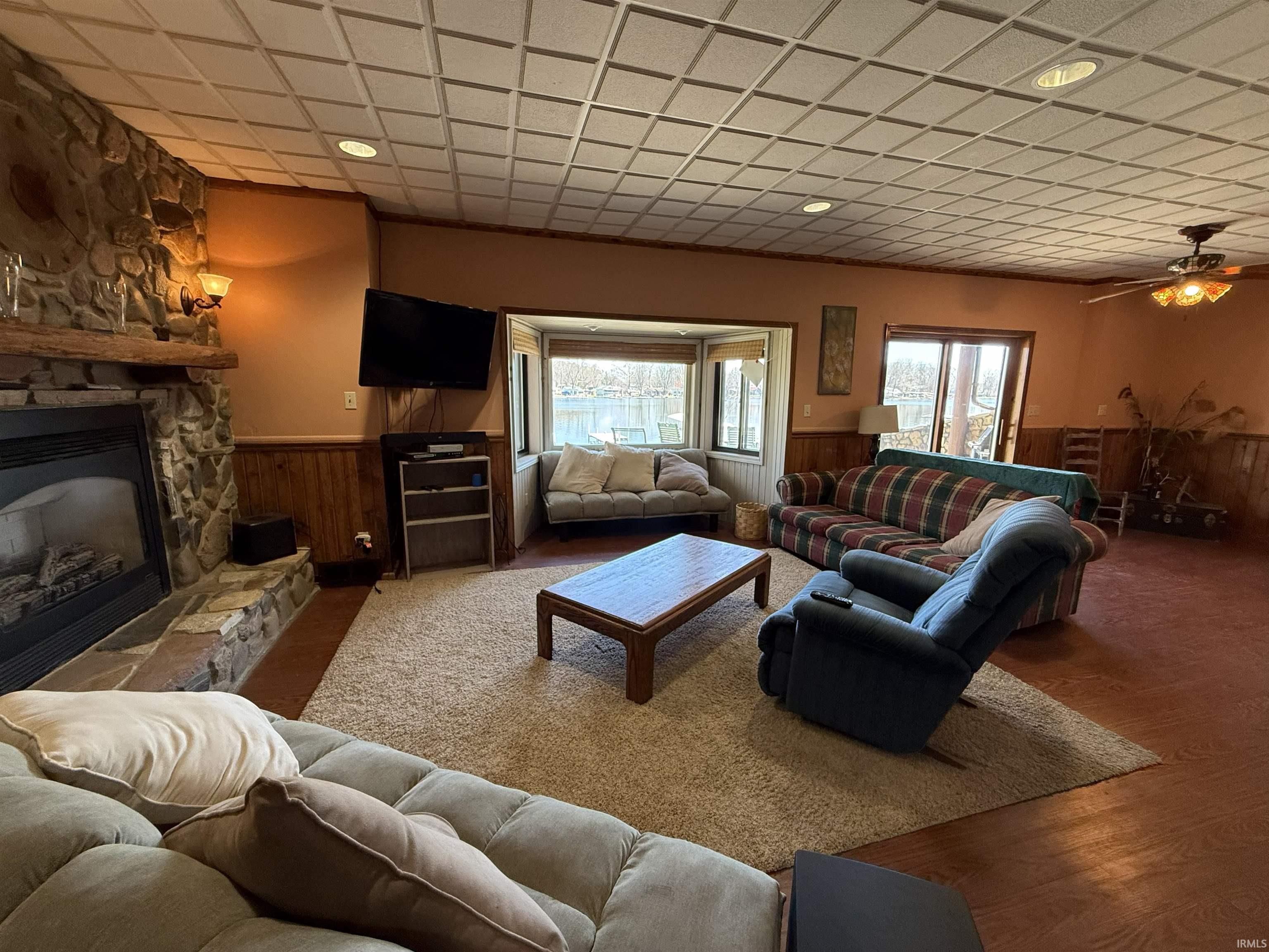 Living room featuring wainscoting, wood walls, a stone fireplace, plenty of natural light, and a ceiling fan
