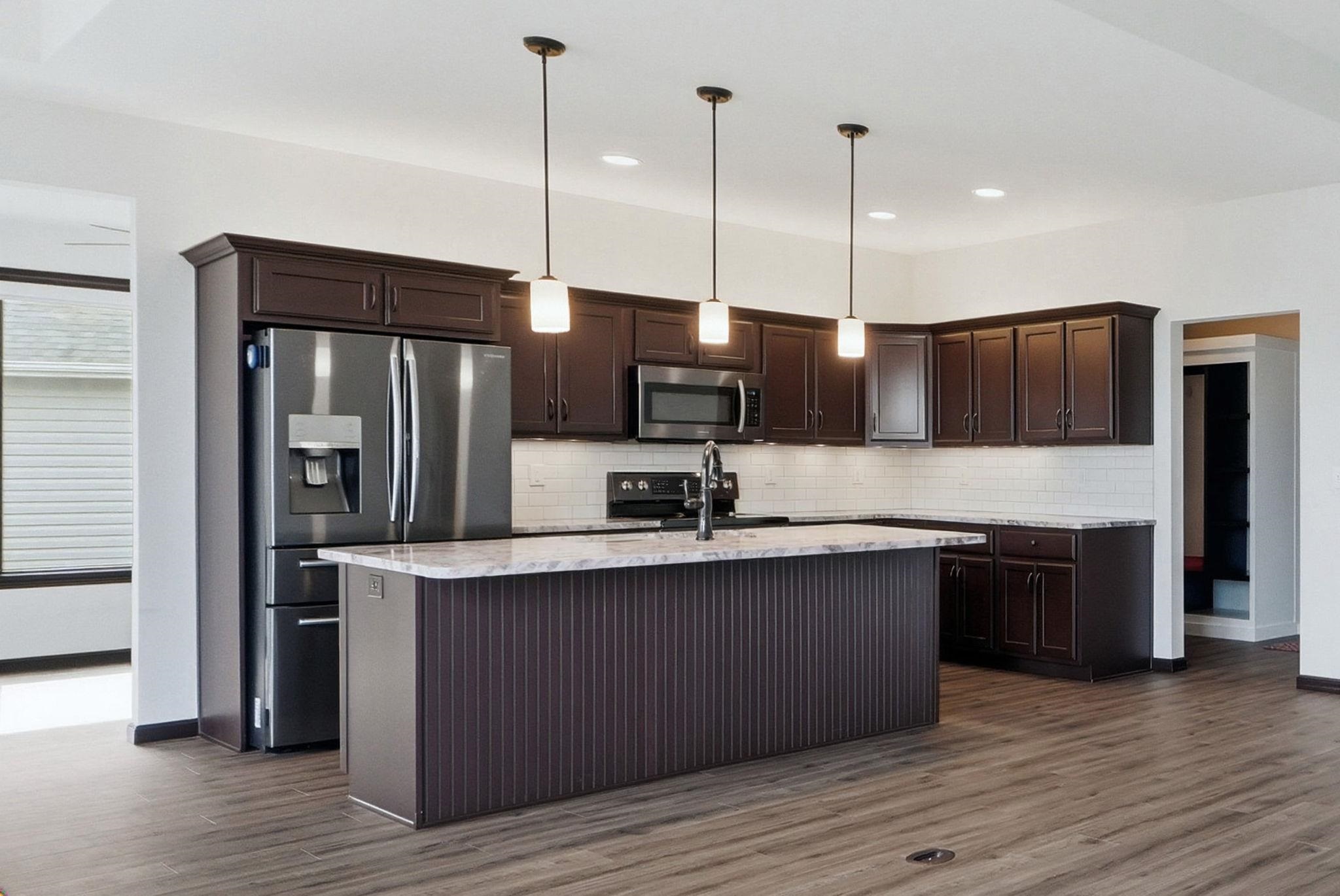 Kitchen featuring dark wood finish cabinetry, stainless steel appliances, a kitchen island with sink, and light stone countertops