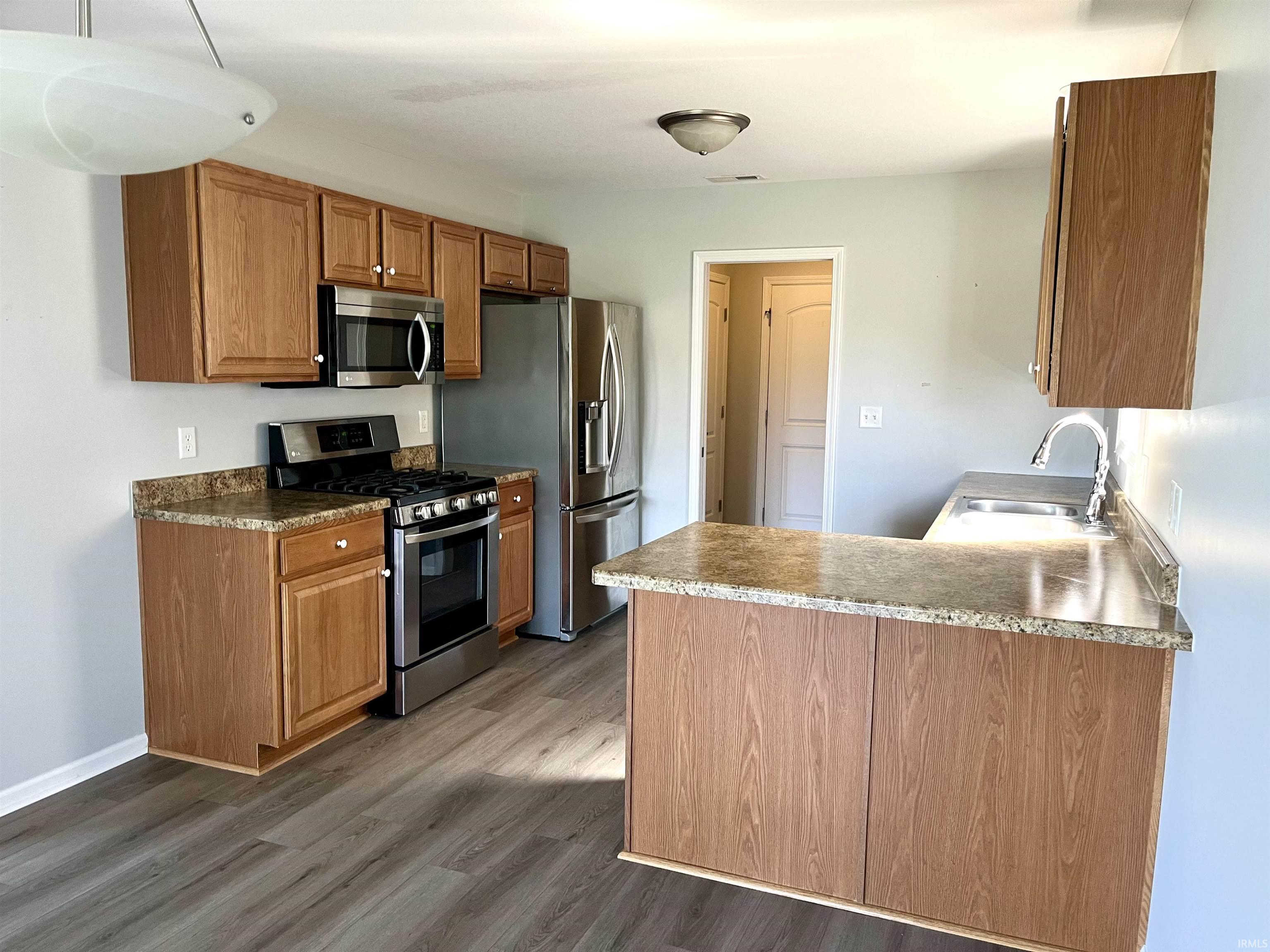 Kitchen featuring stainless steel appliances, a peninsula, wood finish cabinets, dark wood-style flooring, and light countertops
