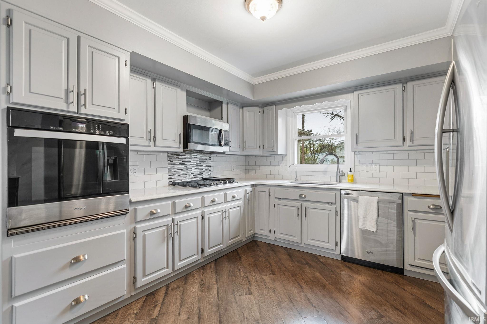 Kitchen with stainless steel appliances, decorative backsplash, dark wood-style floors, crown molding, and light stone counters