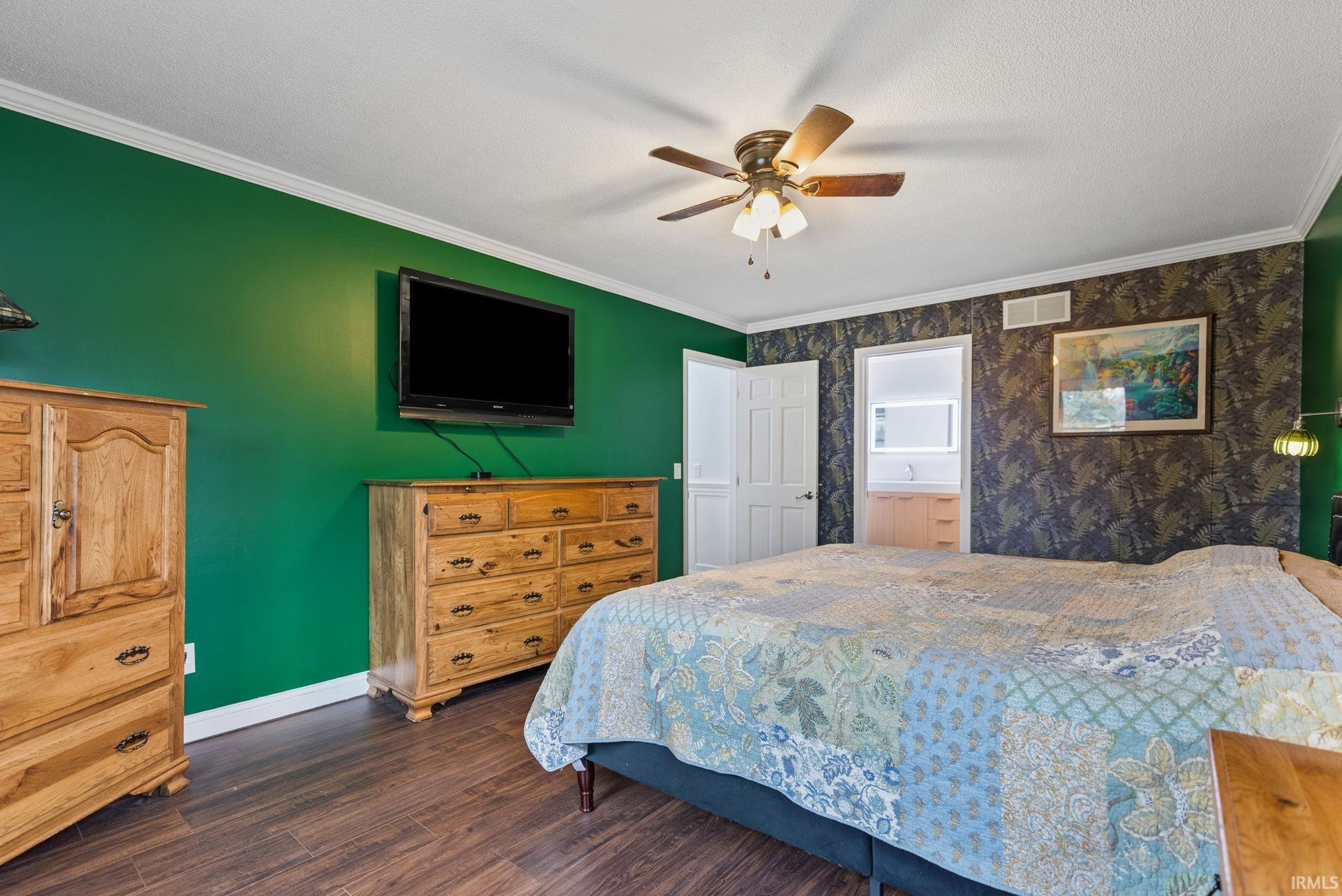 Bedroom with ornamental molding, dark wood-type flooring, a ceiling fan, connected bathroom, and wallpapered walls