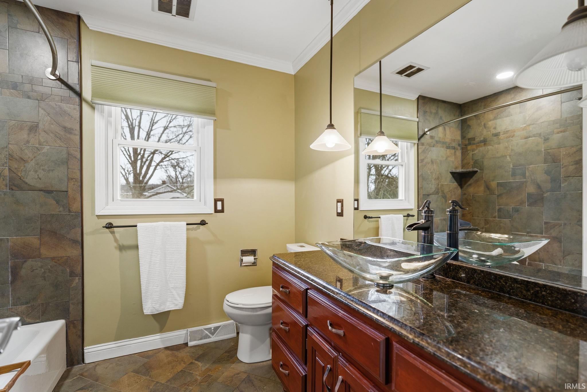 Full bath with dark stone finish floors, vanity, crown molding, healthy amount of natural light, and washtub / shower combination