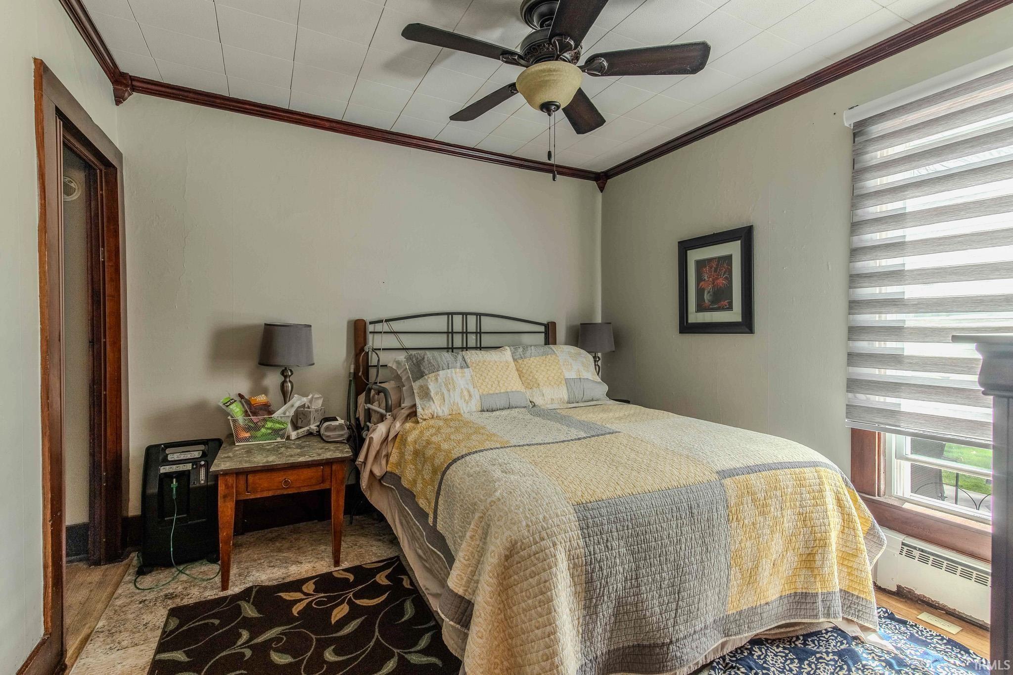 Bedroom with multiple windows, baseboard heating, crown molding, ceiling fan, and wood finished floors