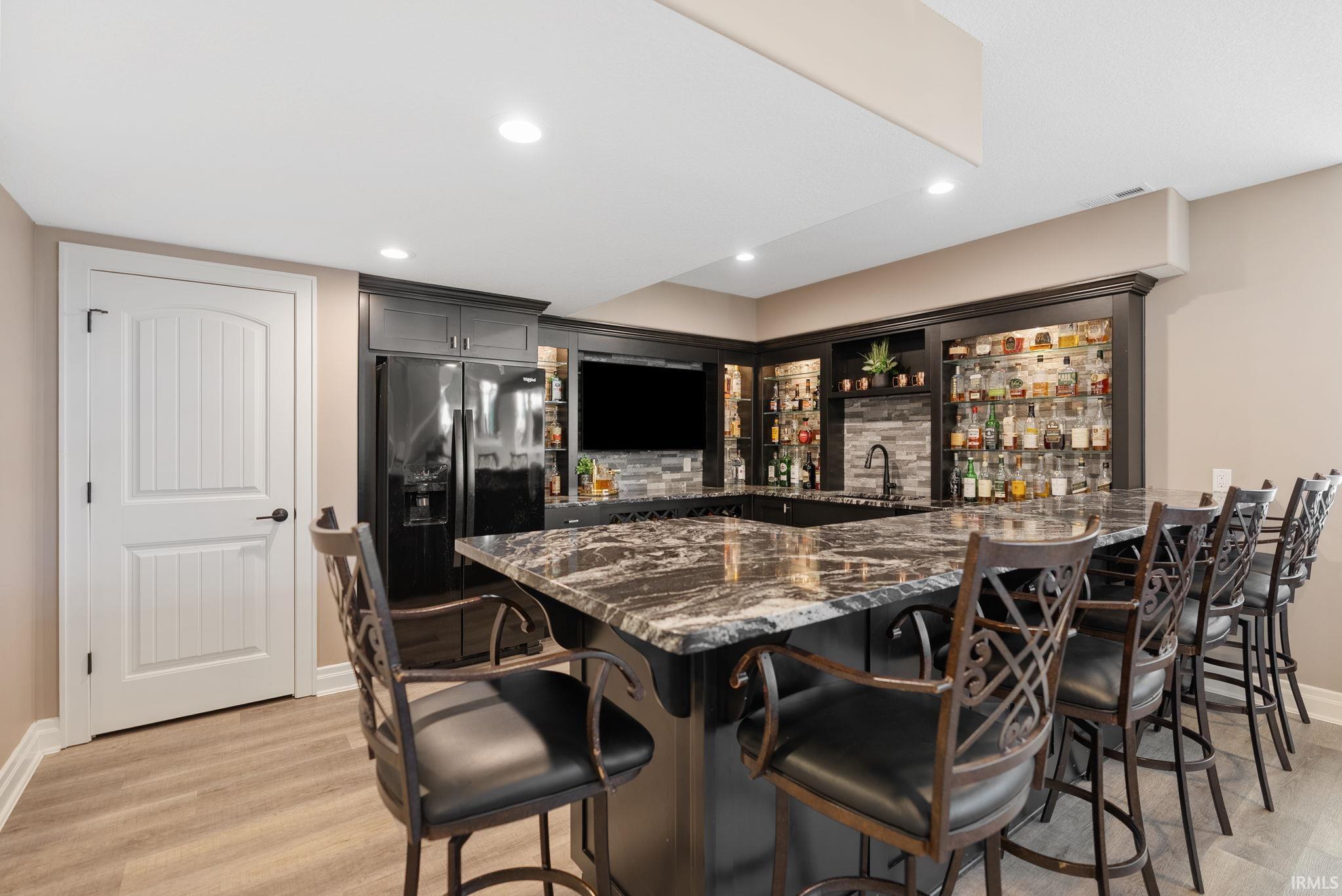 Indoor wet bar featuring dark stone countertops, black refrigerator with ice dispenser, light wood-type flooring, and recessed lighting