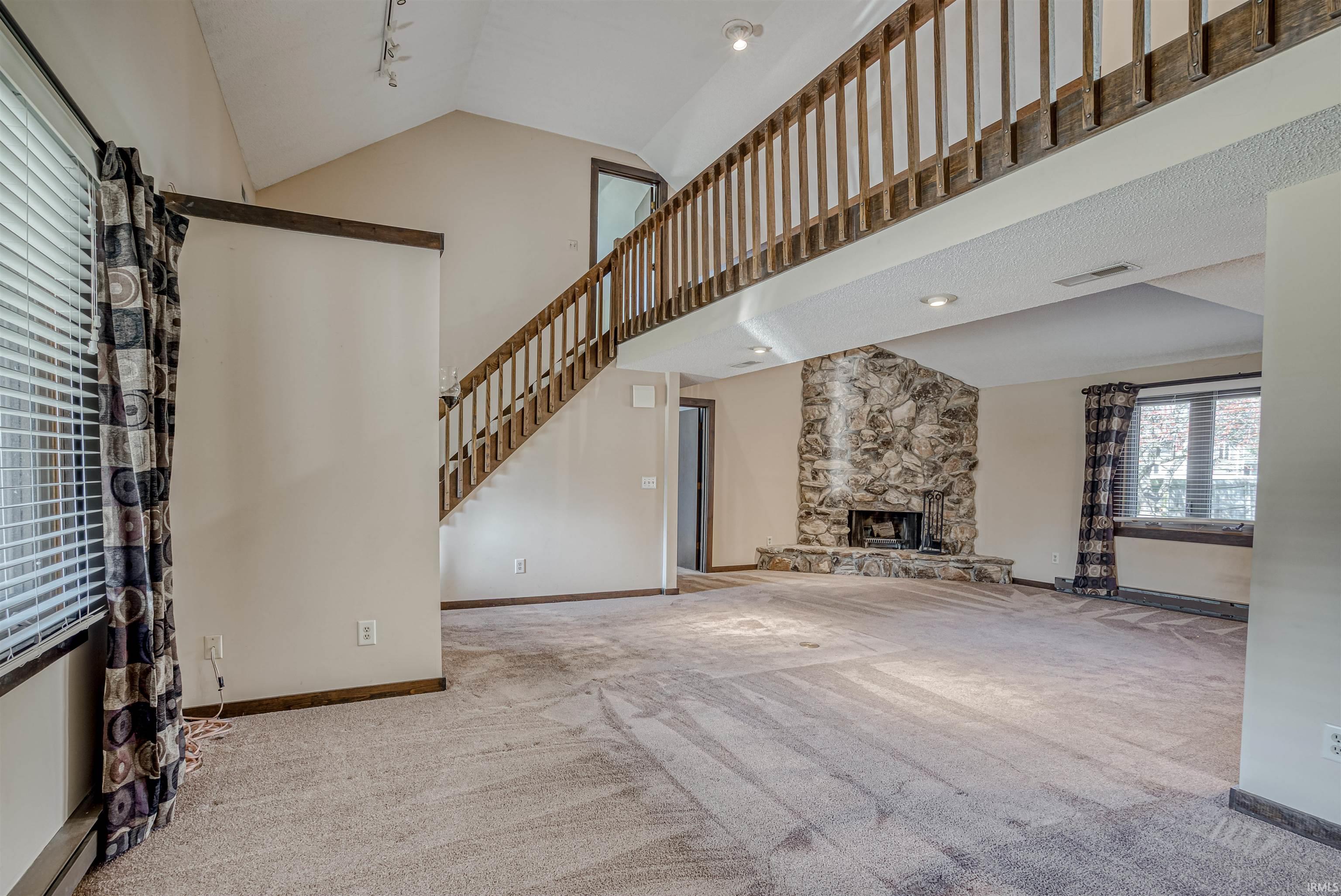 Unfurnished living room featuring track lighting, light colored carpet, vaulted ceiling, and a stone fireplace