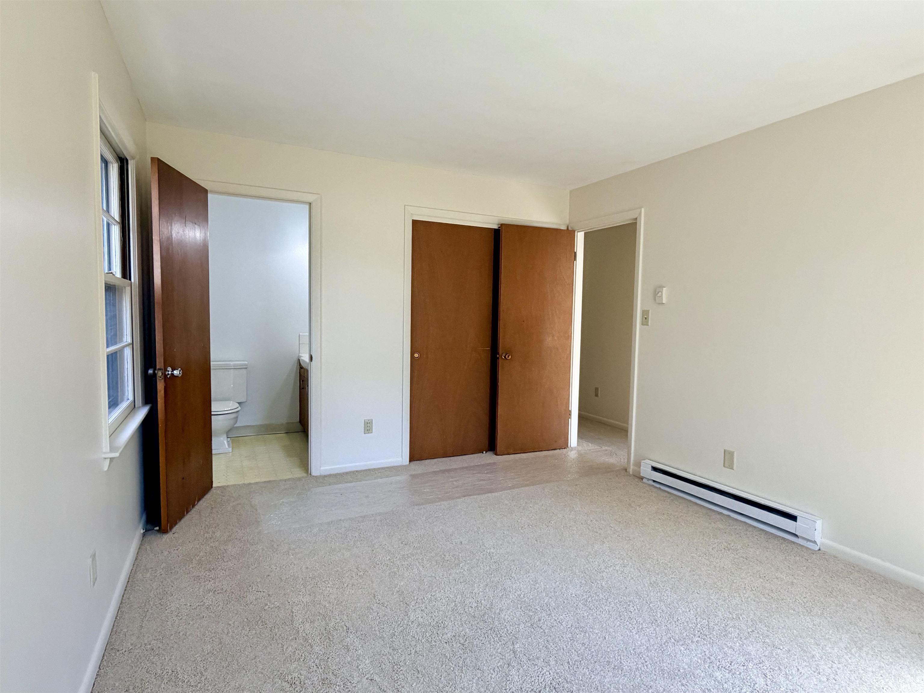 Unfurnished bedroom featuring a baseboard radiator, light colored carpet, connected bathroom, and a closet