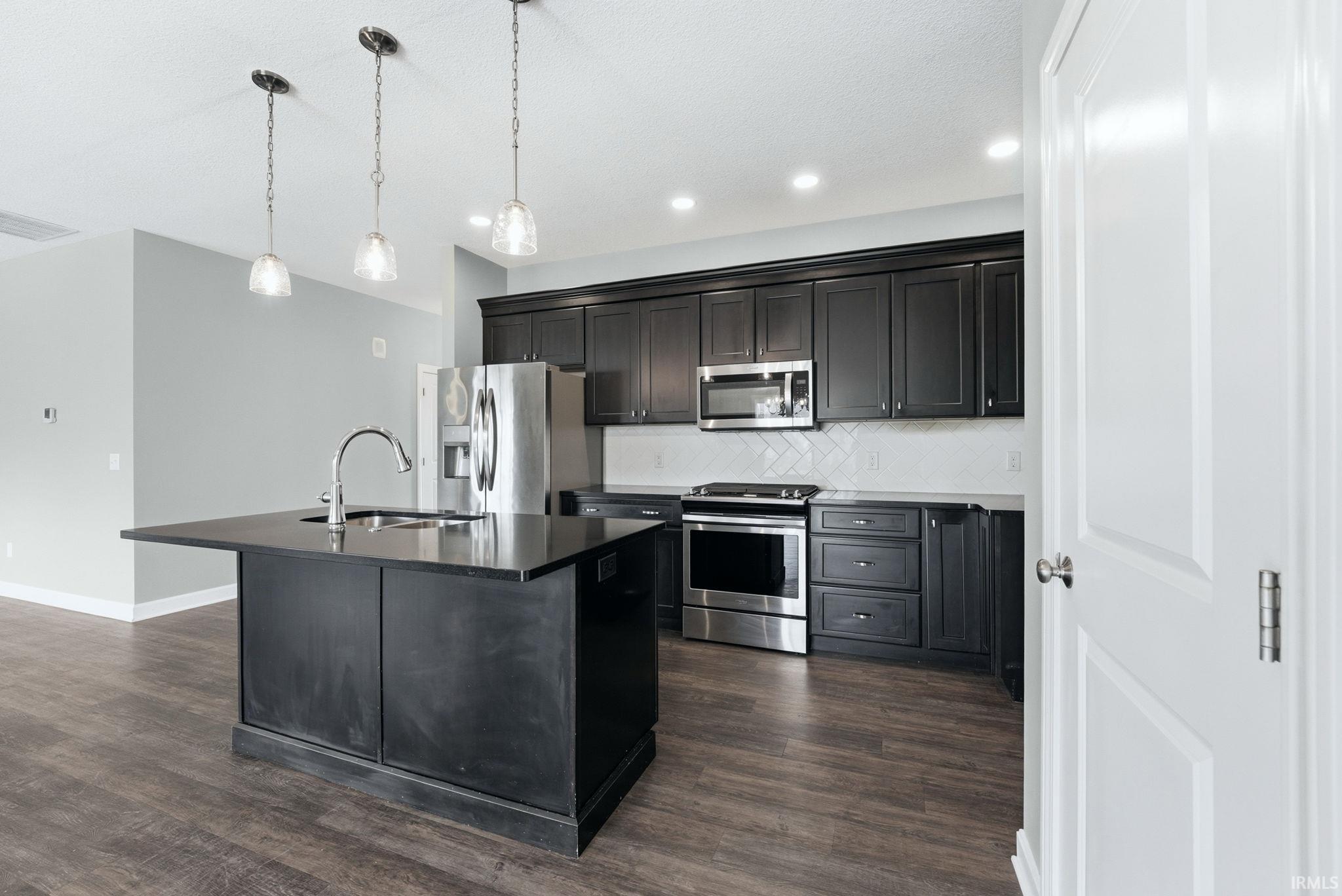 Kitchen with stainless steel appliances, an island with sink, dark wood-style flooring, and hanging light fixtures