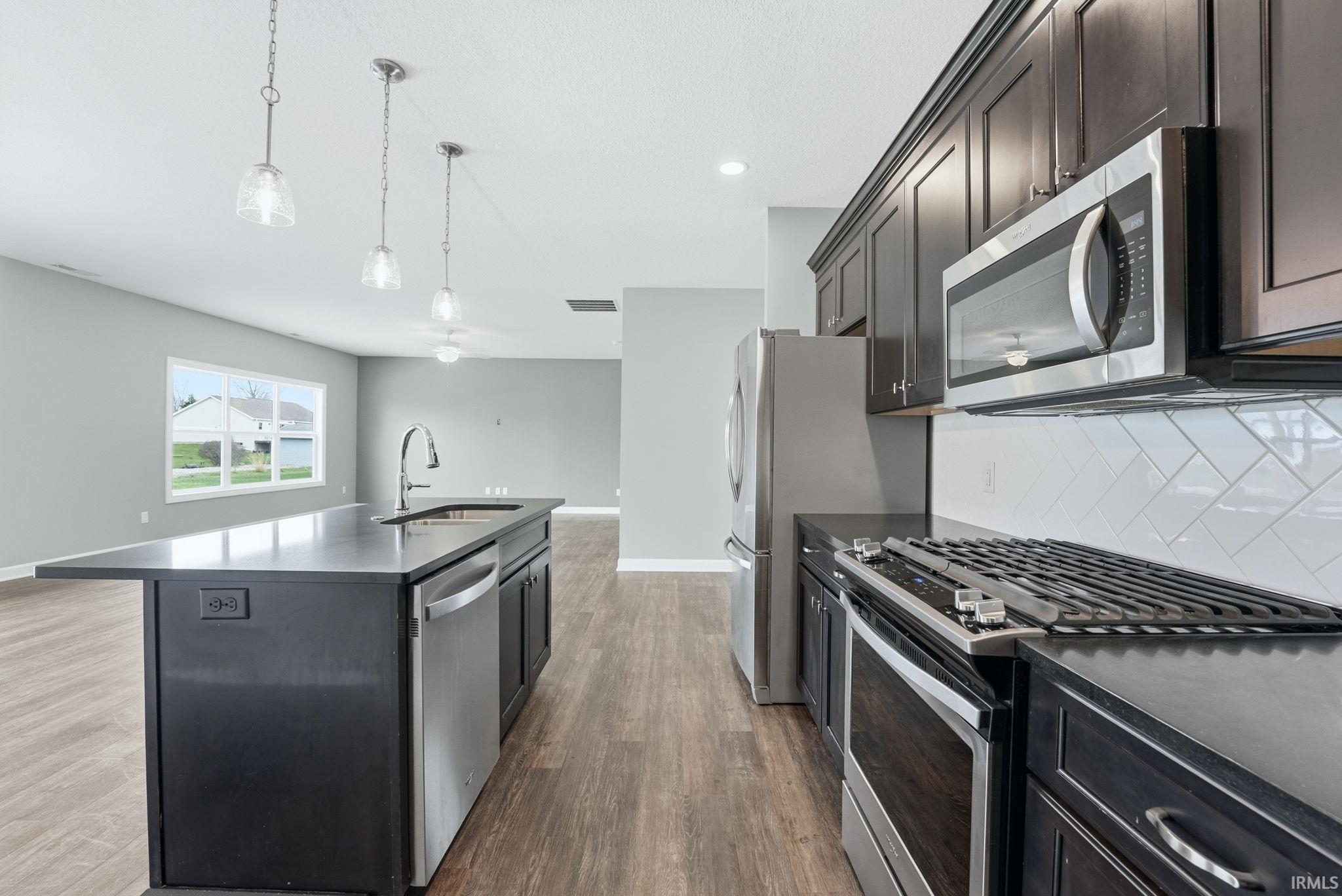 Kitchen featuring stainless steel appliances, pendant lighting, an island with sink, dark stone countertops, and dark wood-type flooring