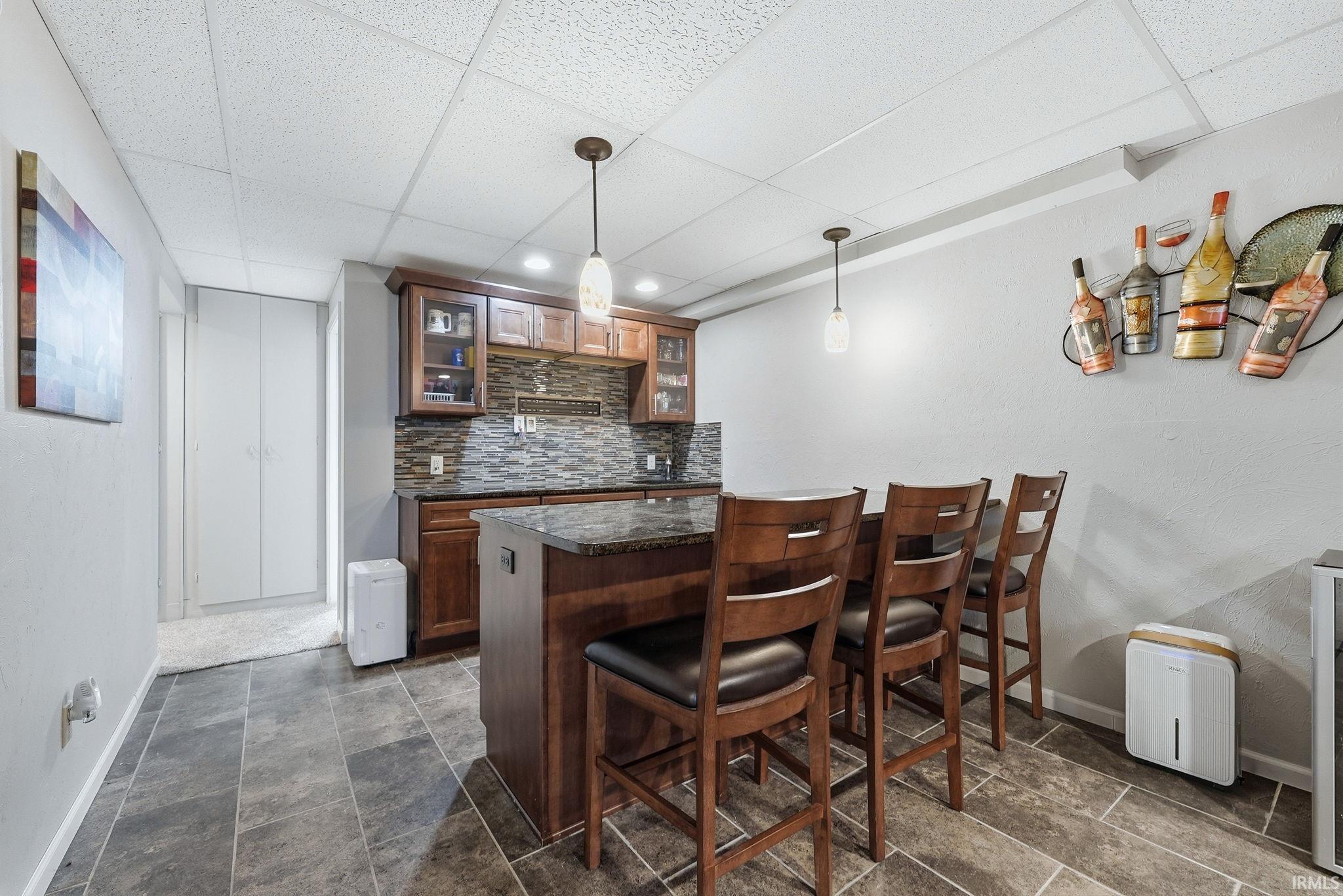 Indoor wet bar with a paneled ceiling, pendant lighting, glass fronted cabinets, backsplash, and dark stone counters
