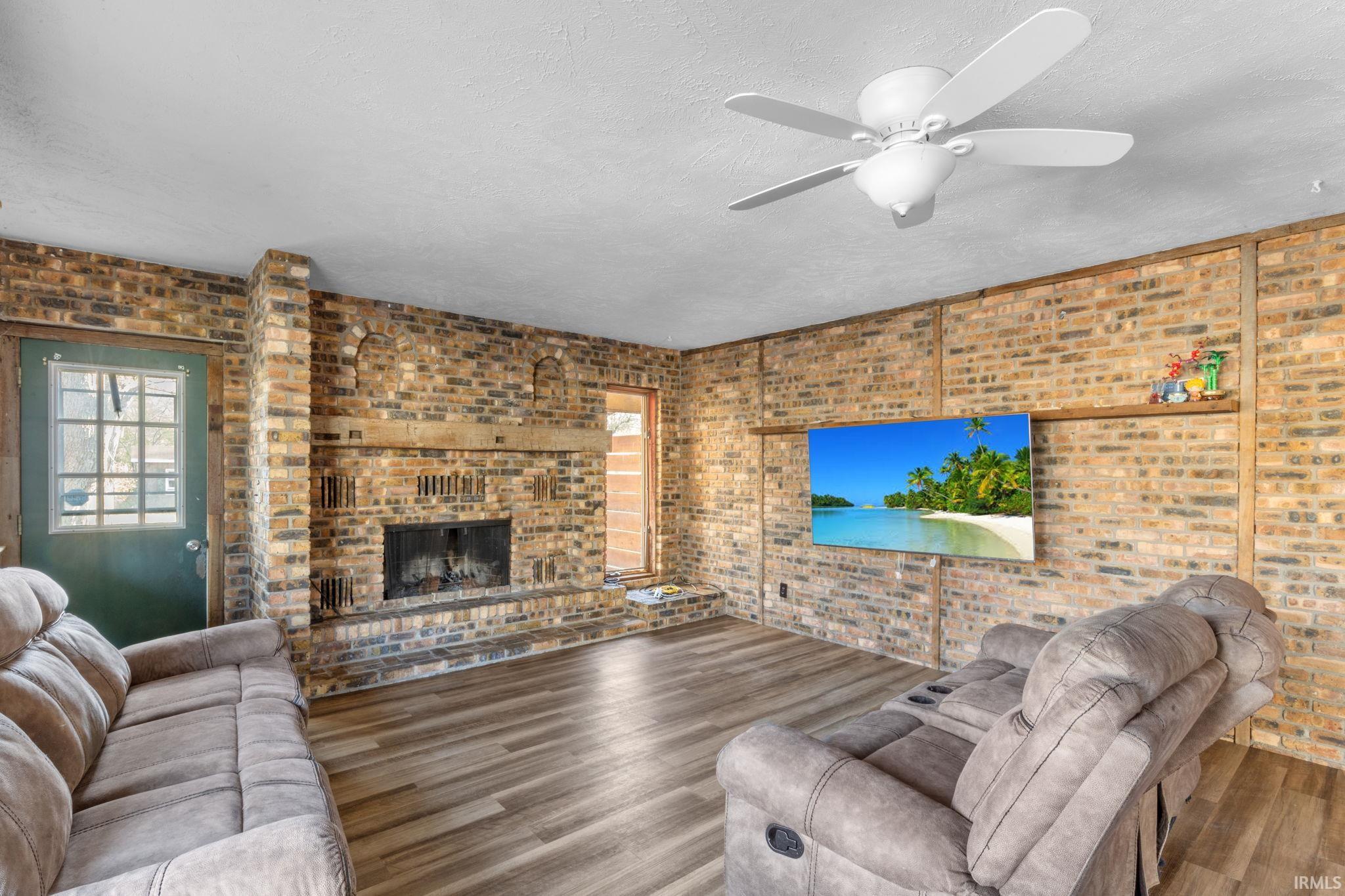 Living area featuring healthy amount of natural light, a brick fireplace, wood finished floors, ceiling fan, and brick wall