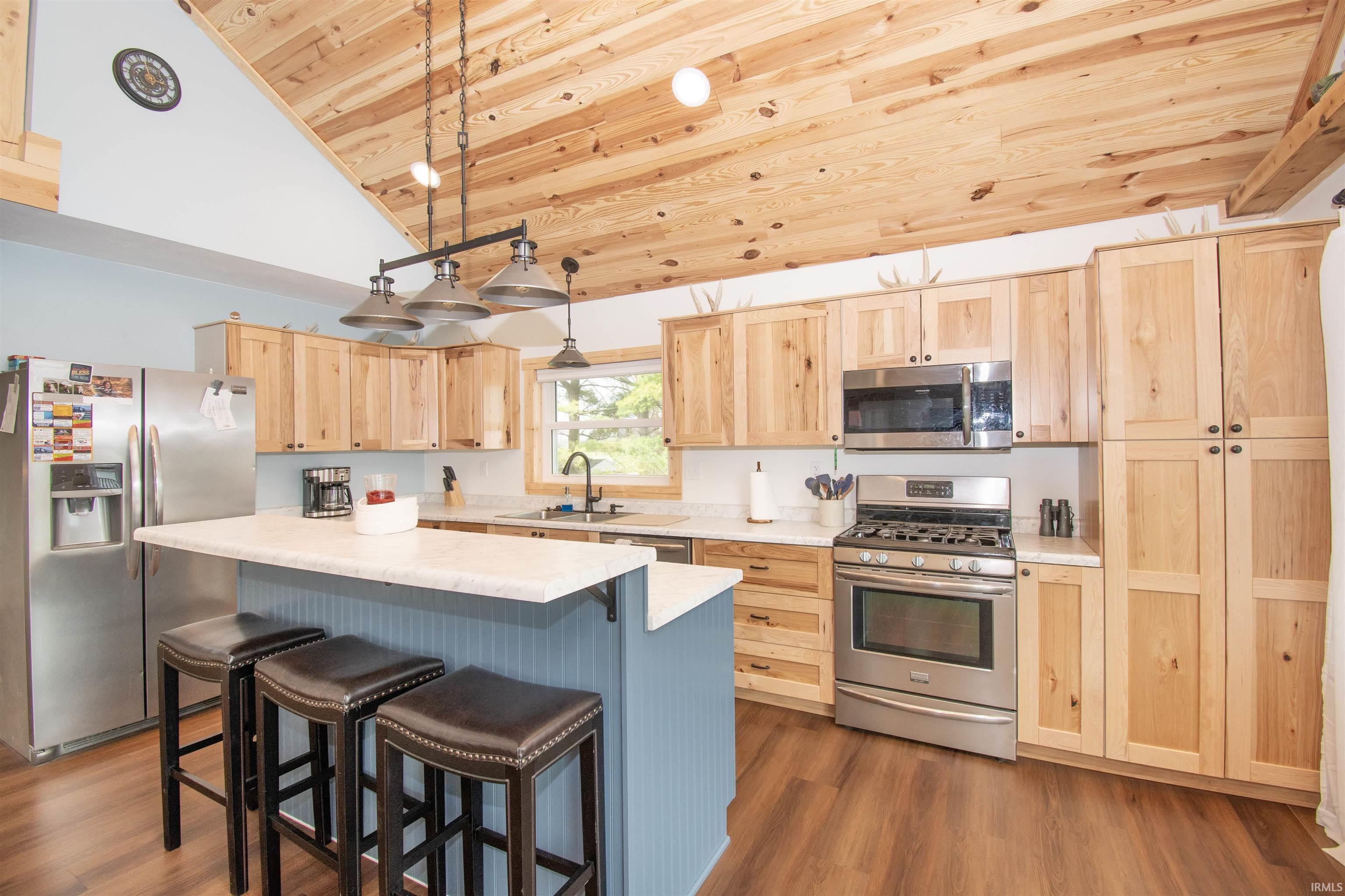 Kitchen with stainless steel appliances, light wood finish cabinetry, dark wood-style floors, a vaulted wooden ceiling, and a breakfast bar