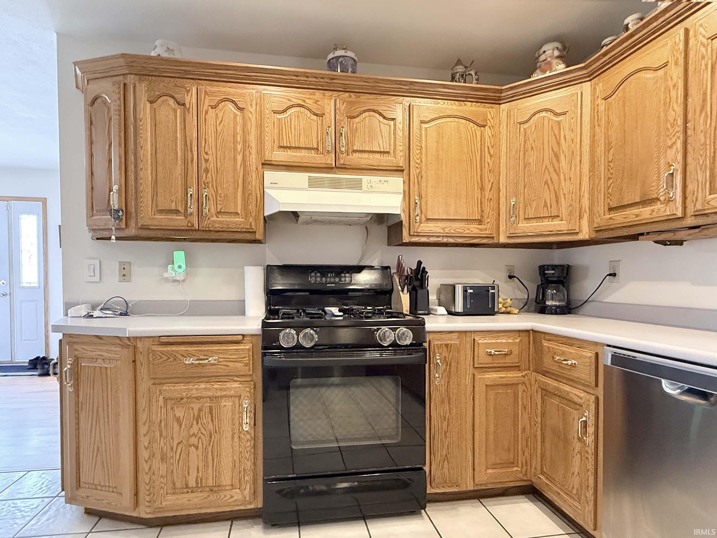 Kitchen with black gas stove, dishwasher, light countertops, light tile patterned floors, and wood finish cabinets